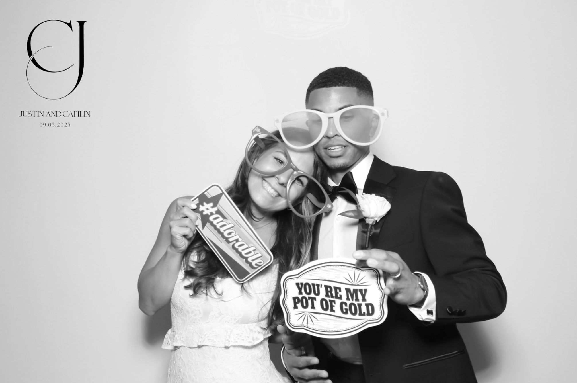 A black-and-white photo of a young couple dressed in wedding attire, wearing large photo booth props including oversized sunglasses. The woman holds up a sign that says '#audiobable' and is leaning into the man, who is in a tuxedo. The background features text on the left side that reads 'CJ' and 'Justin and Caitlin 09.05.2025', indicating a wedding date.