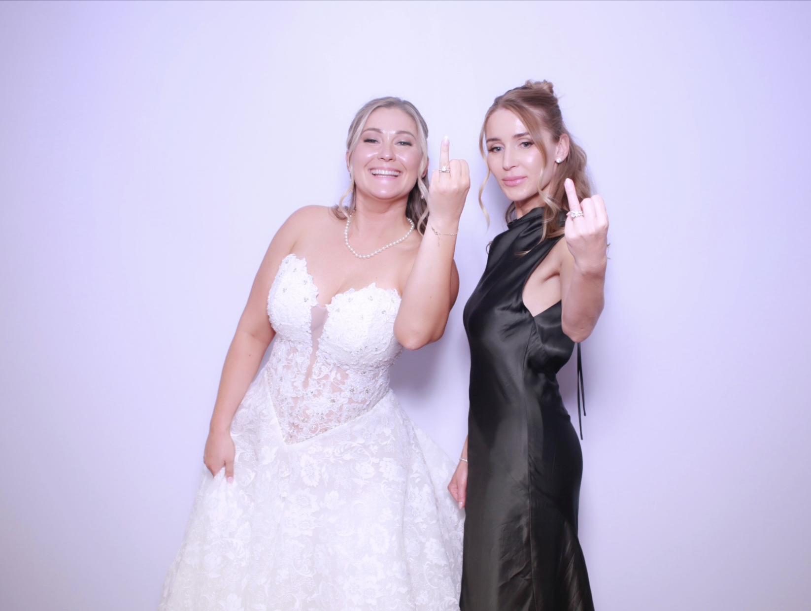 Two women, one in a wedding dress and the other in a black dress, standing against a plain white wall, showing middle fingers.