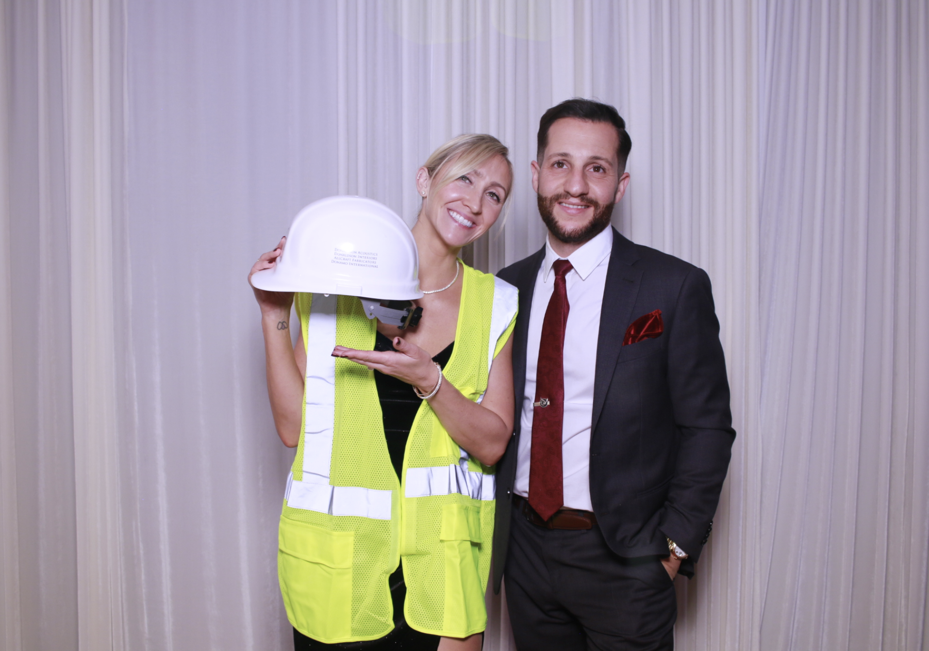 A woman in a yellow safety vest holding a white hard hat stands next to a man in a black suit, white shirt, and red tie; both are smiling in front of a white curtain.