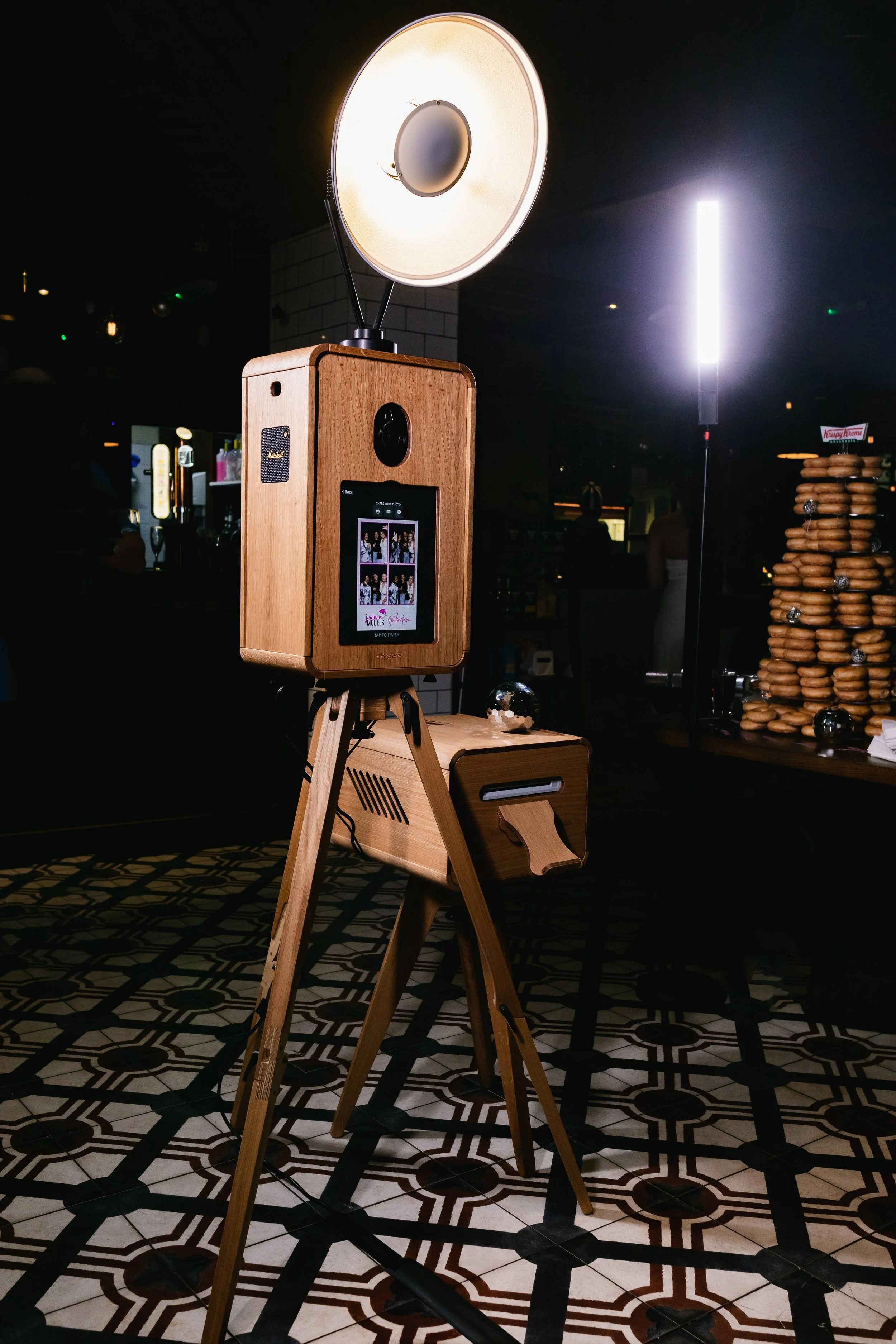 Photo of a photo booth with a wooden exterior, a circular light above it, and a screen displaying photo strips. The display shows a group of people taking pictures. The scene is indoors with decorative patterned flooring and a stack of bagels on the 