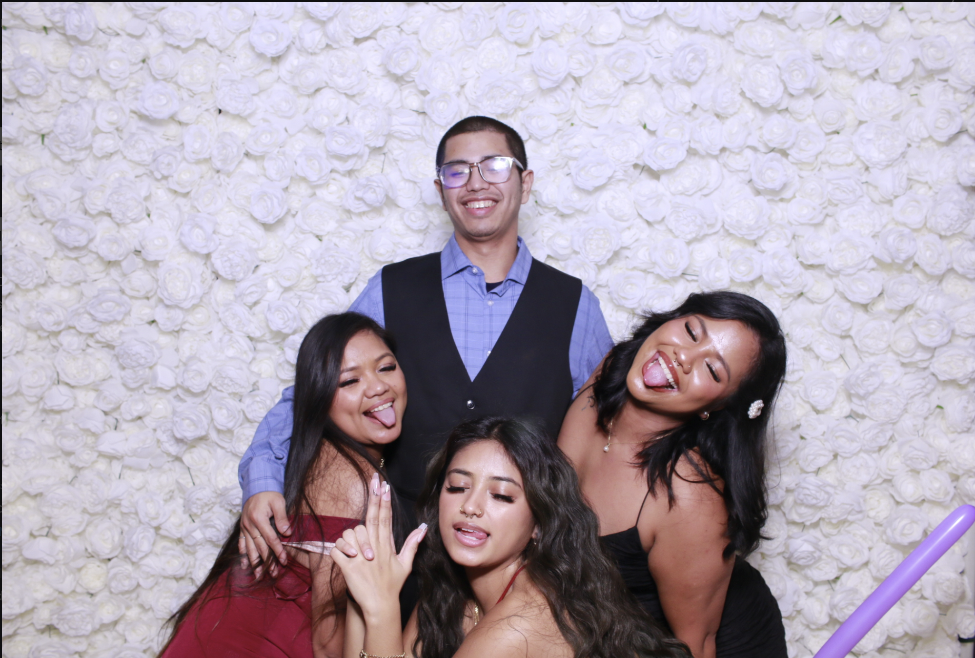 Group of people posing playfully in front of a wall of white roses at a celebration or event.