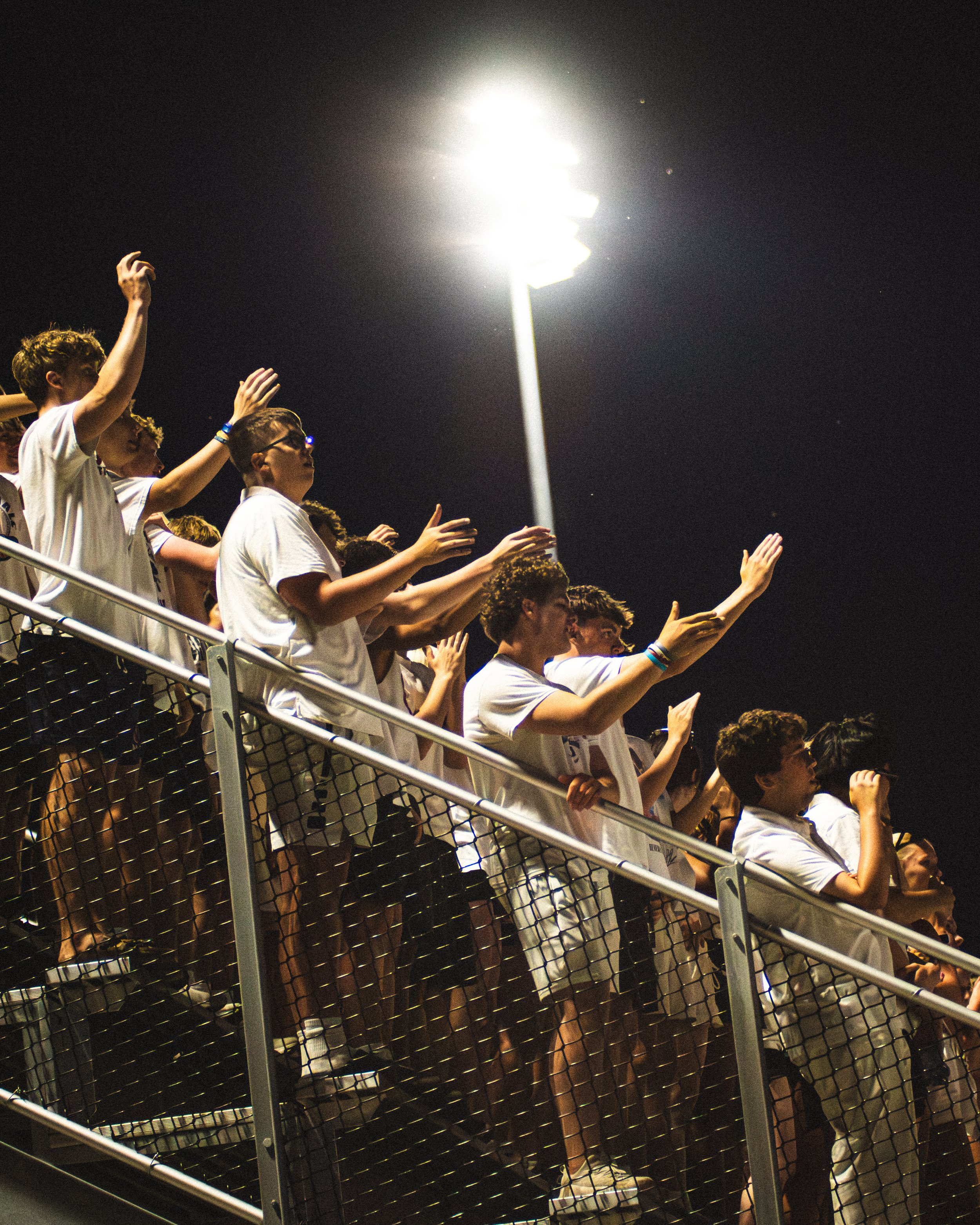 A group of young people standing by a fence at night, raising their hands, under bright stadium lights.