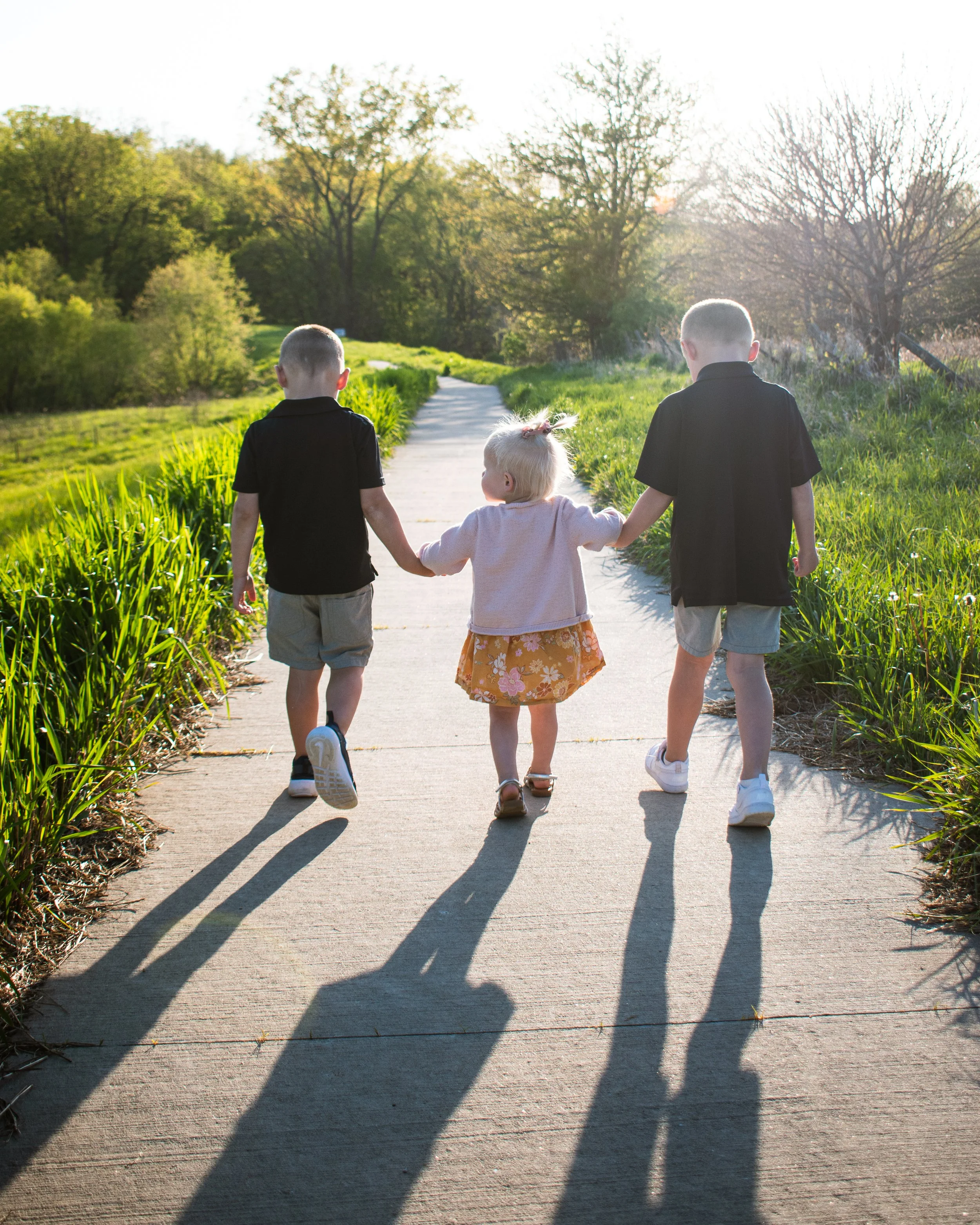 Three children walking on a sidewalk during sunset, holding hands with shadows cast on the ground, lush greenery on both sides, trees in the background, and a bright sky.