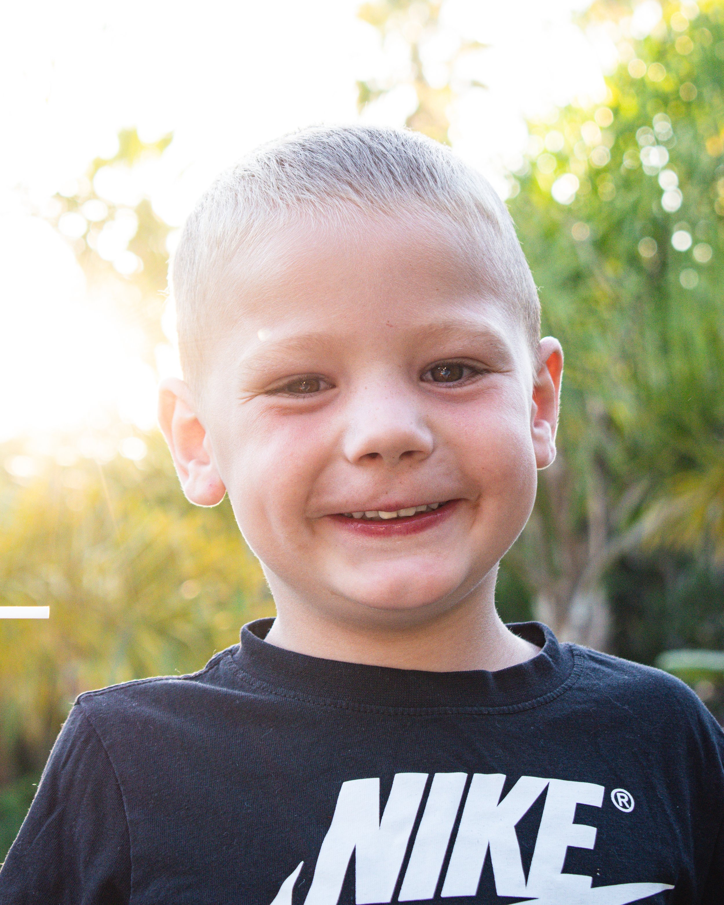 Close-up of a young boy with short blond hair and brown eyes outdoors, smiling slightly, wearing a black Nike shirt, with blurred green trees in the background.
