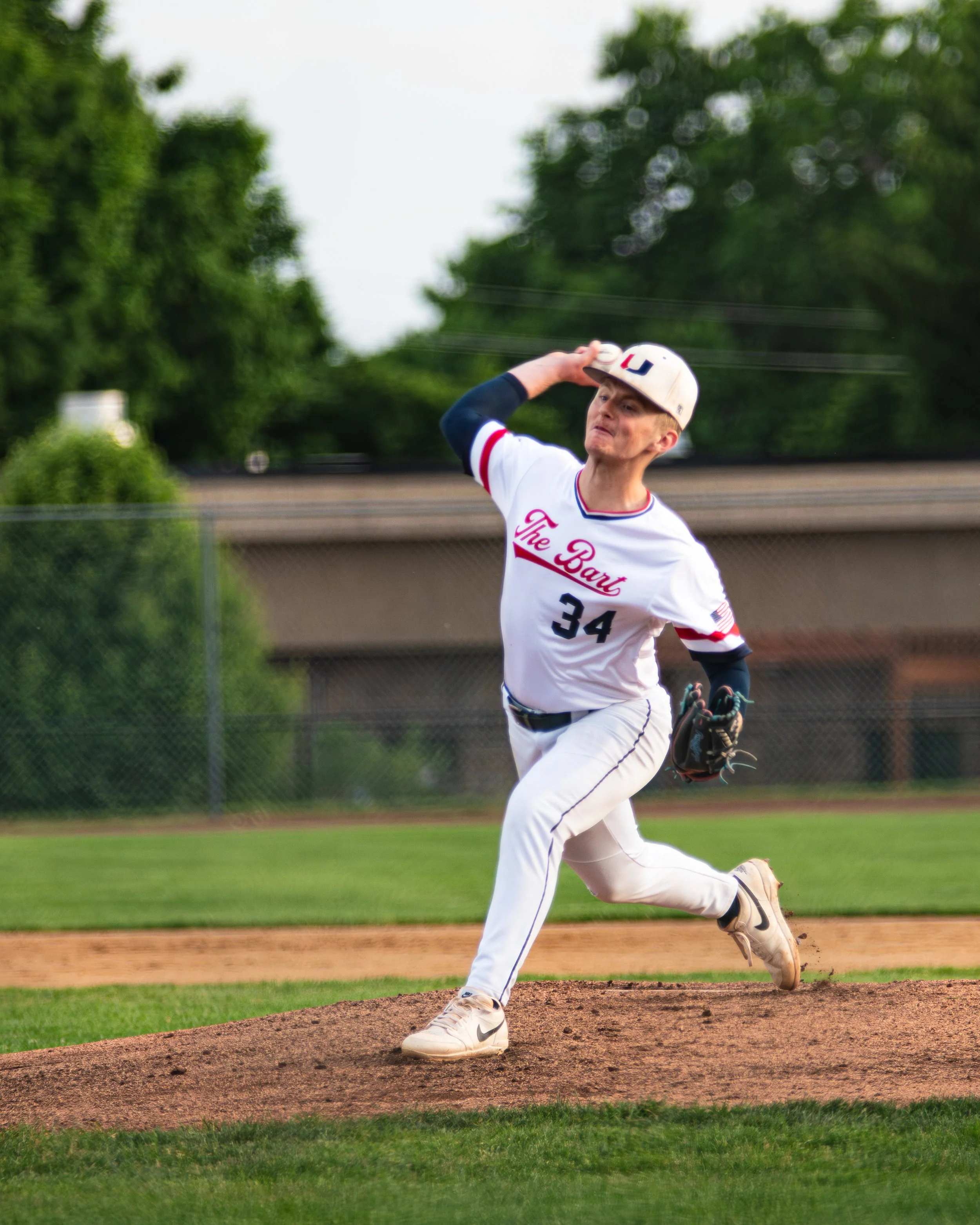 A young baseball player in a white uniform with red and blue trim is running on the pitcher's mound during a game, with a baseball glove on his left hand and wearing a cap with the letter 'S' on it, on a baseball field with a grassy background and trees.