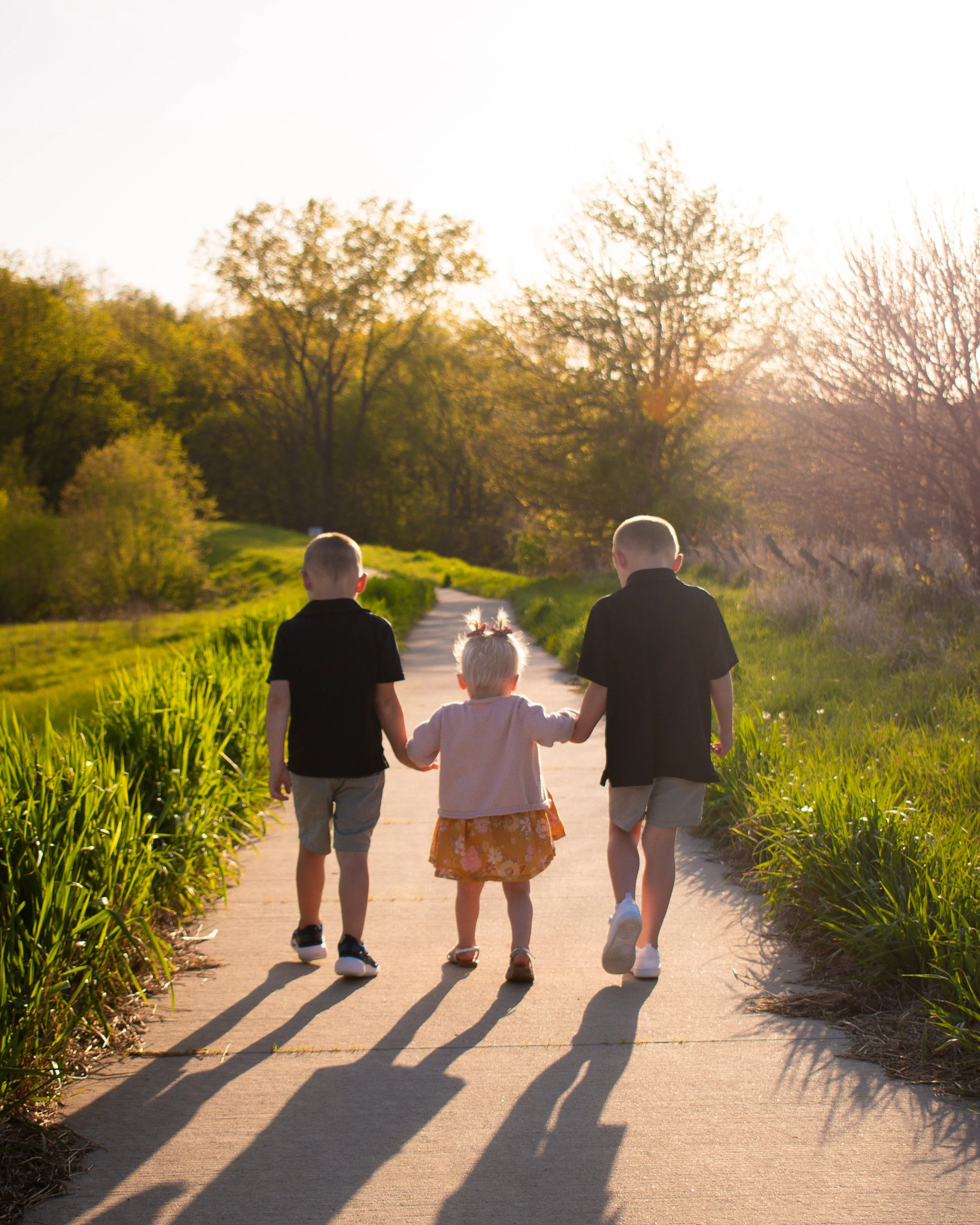 Four children holding hands and walking down a sunny sidewalk surrounded by green trees and tall grass, with long shadows cast on the ground.