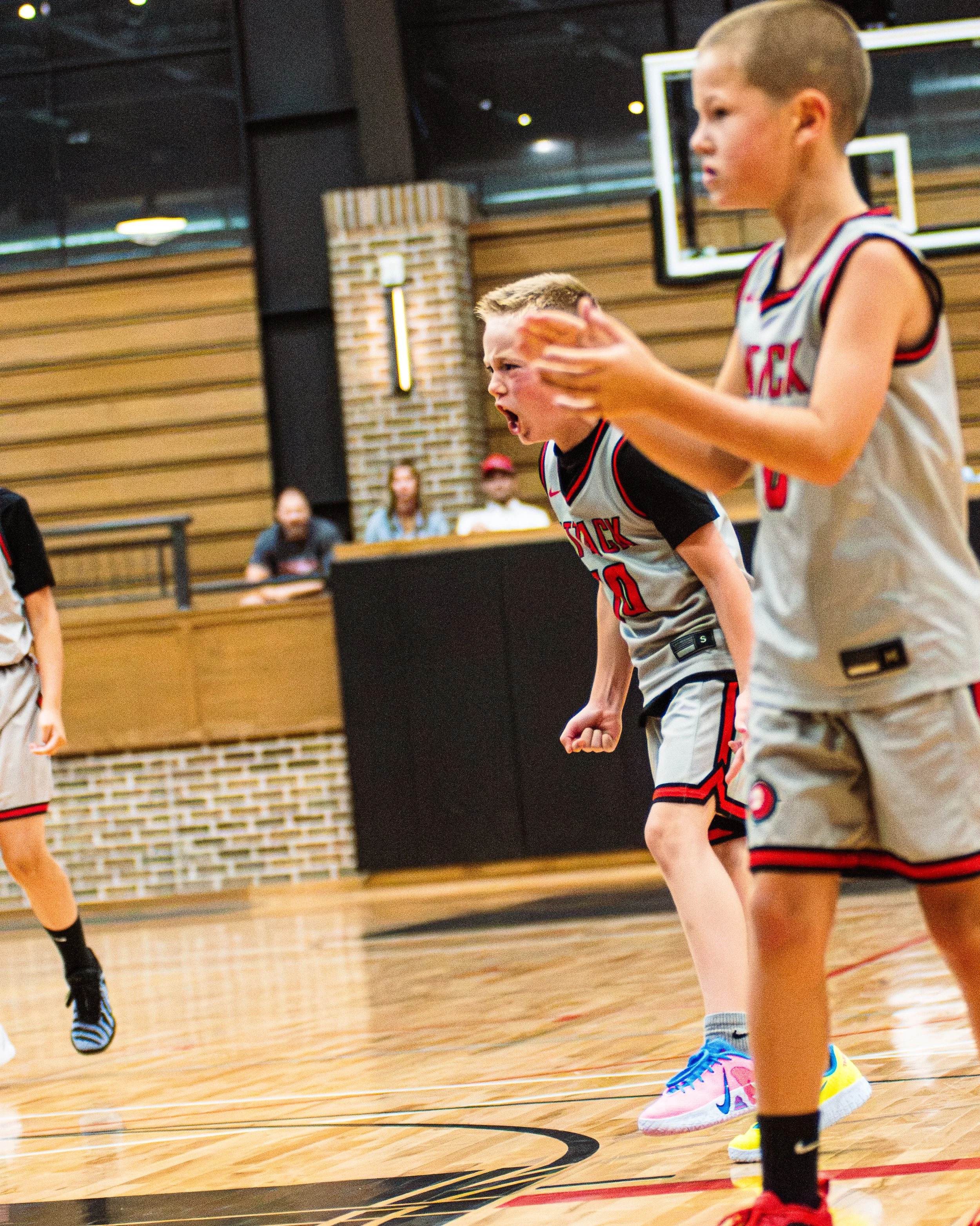Young boys playing basketball in an indoor gymnasium with wooden flooring, wearing basketball uniforms and sneakers.