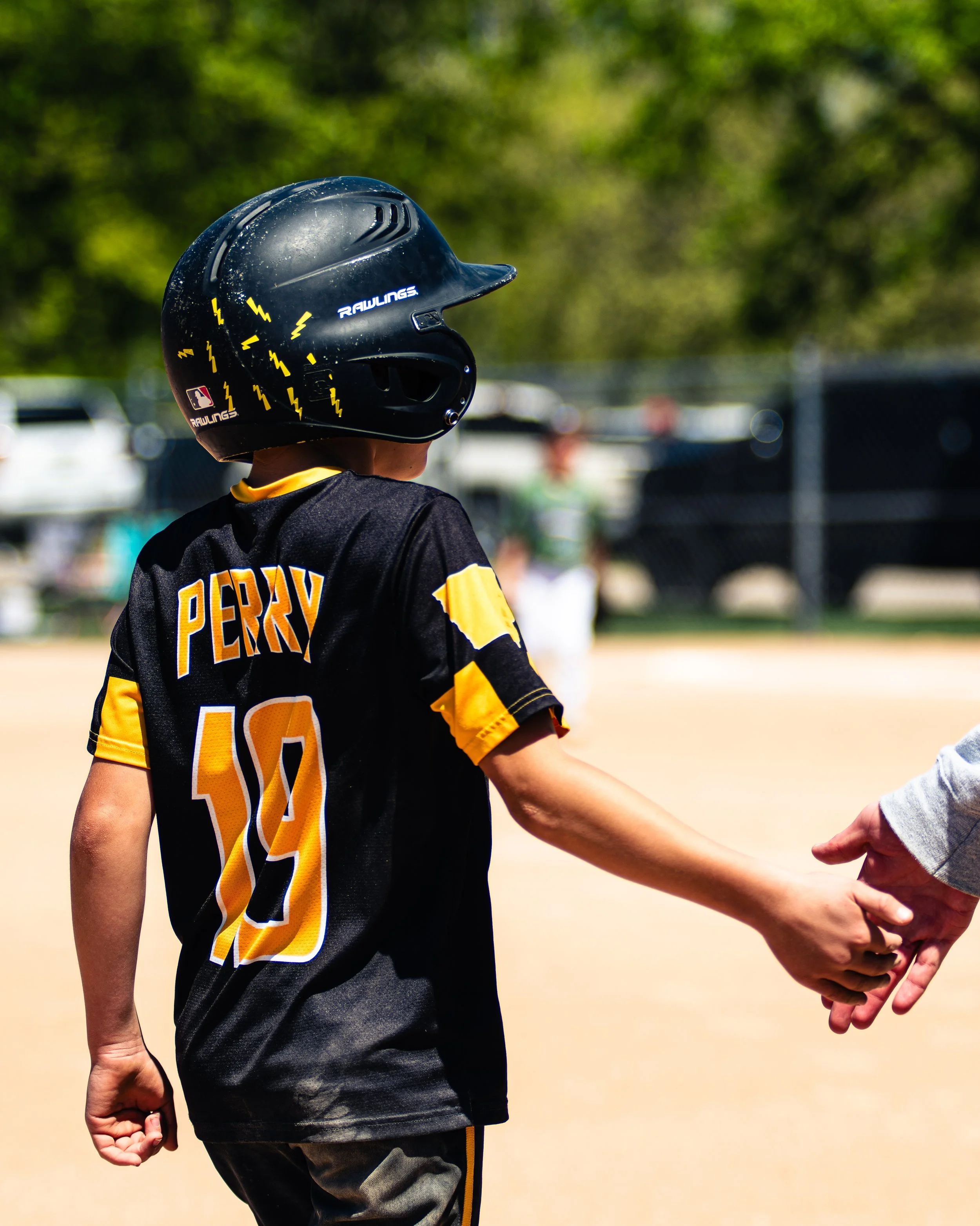 A young baseball player wearing a black helmet and team uniform holding hands with another person at a baseball field.