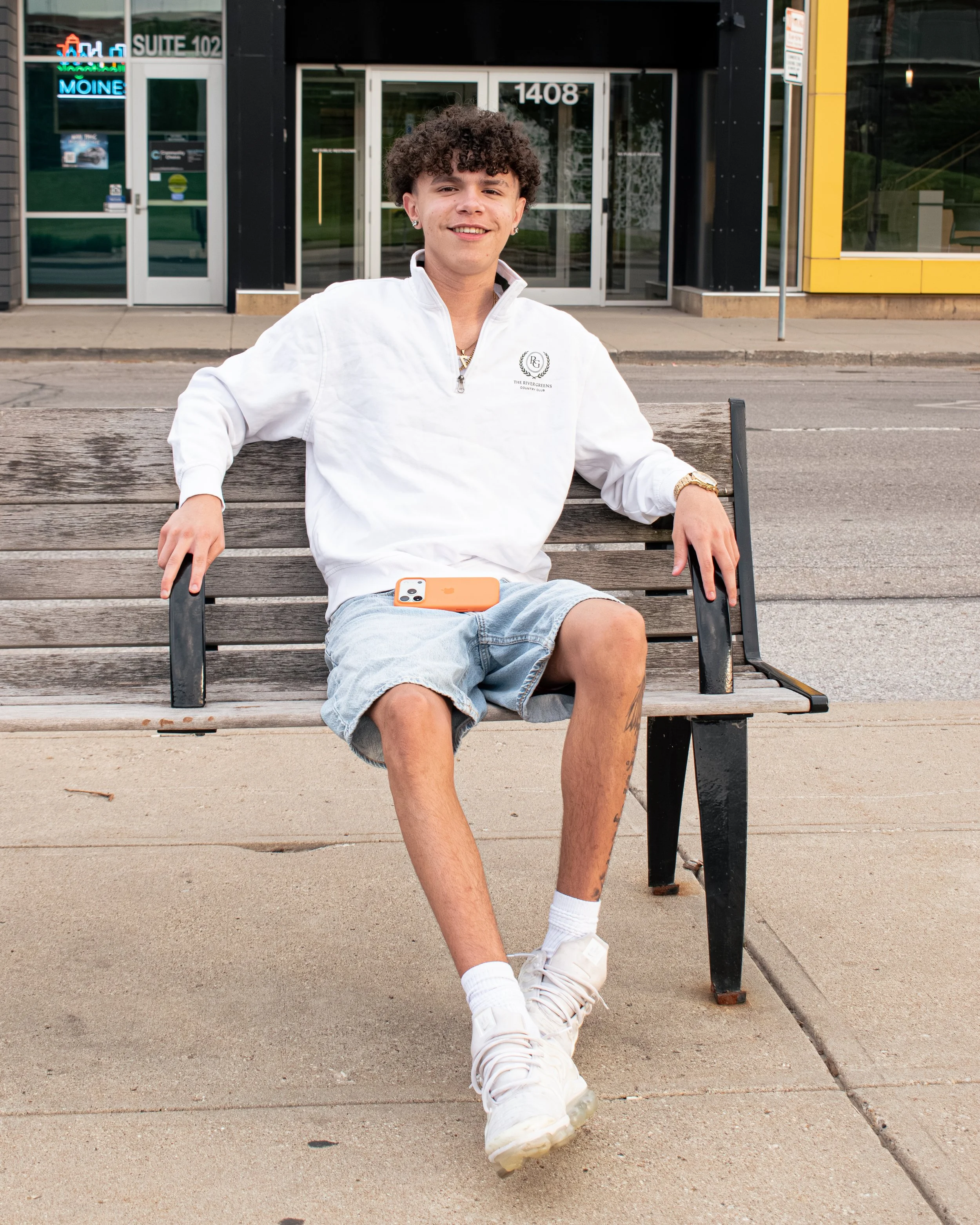 Young man with curly hair sitting on a wooden bench outdoors in front of a building with glass doors and colorful storefront signs, wearing a white quarter-zip pullover, denim shorts, white sneakers, and gold jewelry, smiling at the camera