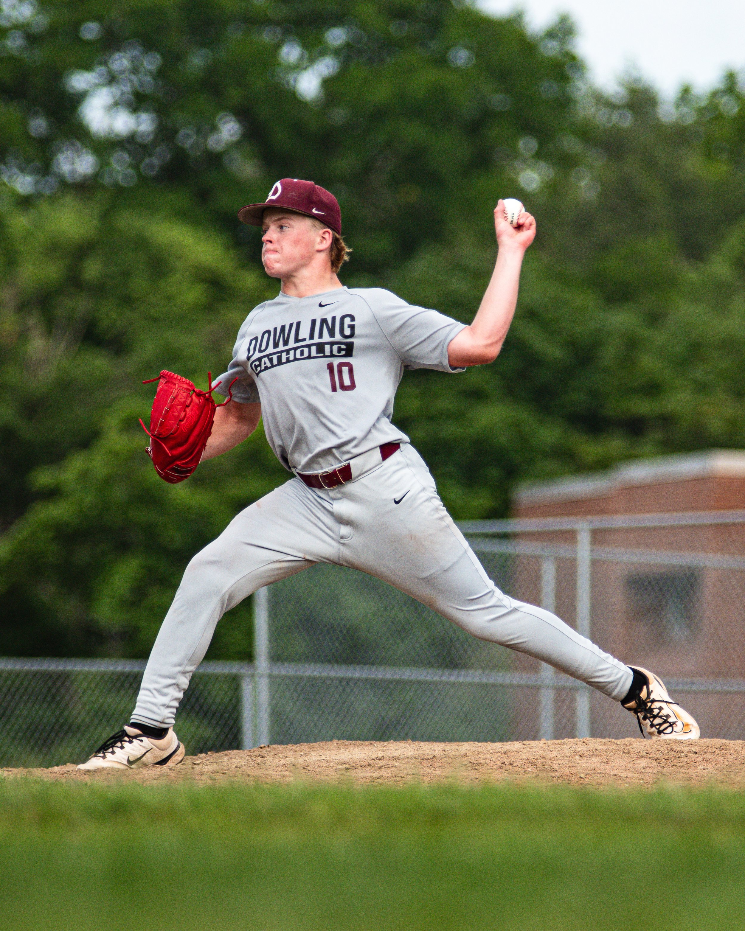 A young baseball pitcher in a gray uniform with maroon and white accents, throwing a pitch on a baseball field. The pitcher is in a stance with his right arm raised, holding a baseball, and his left arm extended downward. He is wearing a maroon cap and white cleats. The background shows blurred green trees and a chain-link fence.