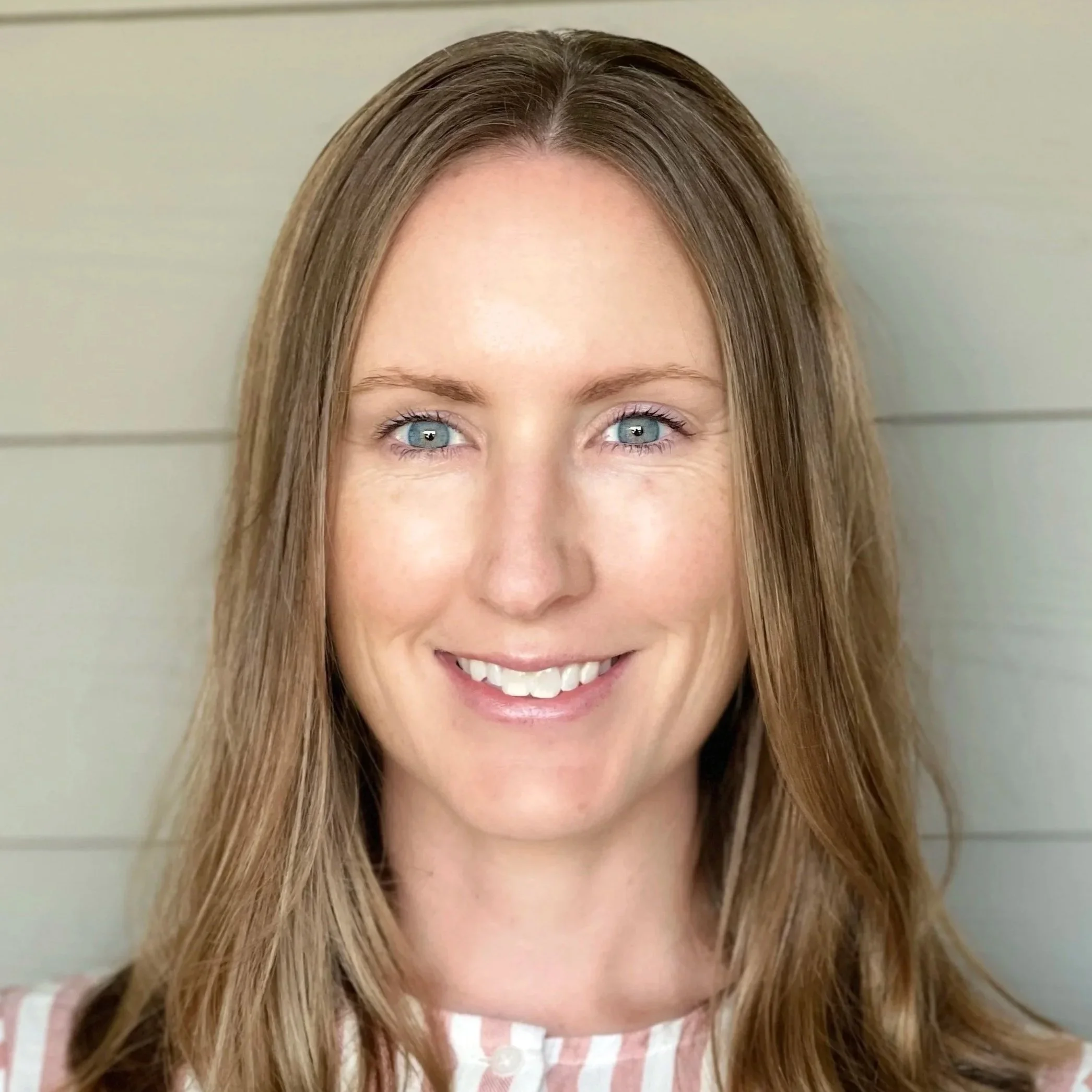 A close-up of a smiling woman with blue eyes and straight light brown hair, wearing a striped top.