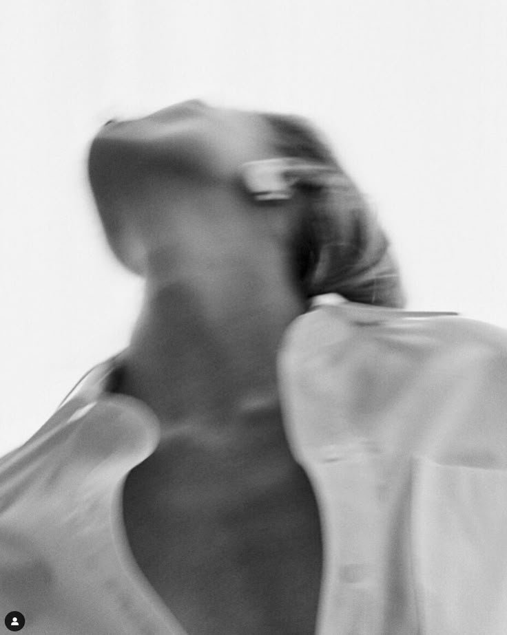 A blurred black and white photo of a person with short hair, wearing glasses, looking upward or sideways. The person is wearing a light-colored top and the background is plain and light-colored.
