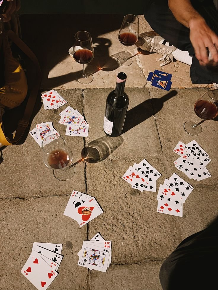 Cards, wine glasses, and a bottle of red wine on an outdoor stone surface, with a person partially visible in the upper right corner.