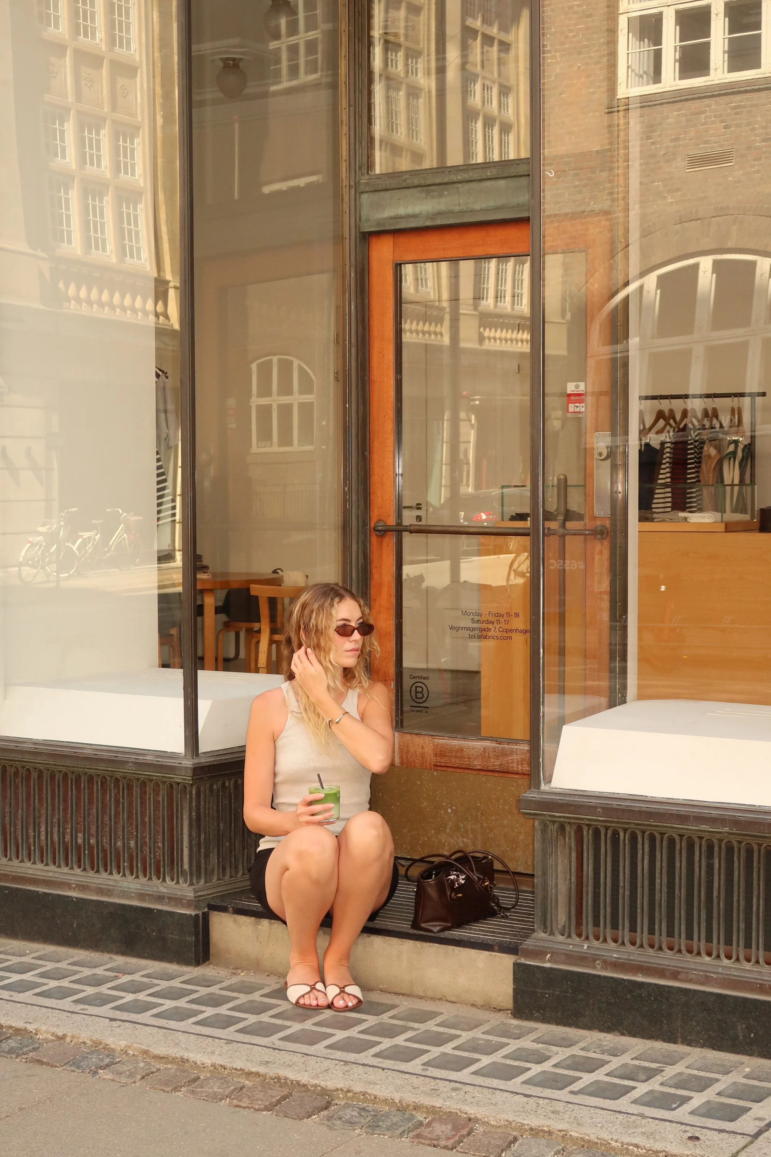 A young woman with blonde hair and sunglasses sitting outside a café, holding a green drink. She is wearing a sleeveless top, shorts, and sandals, with her legs crossed and a brown handbag next to her. The café has a large glass window and door with wooden accents, displaying clothing racks inside.