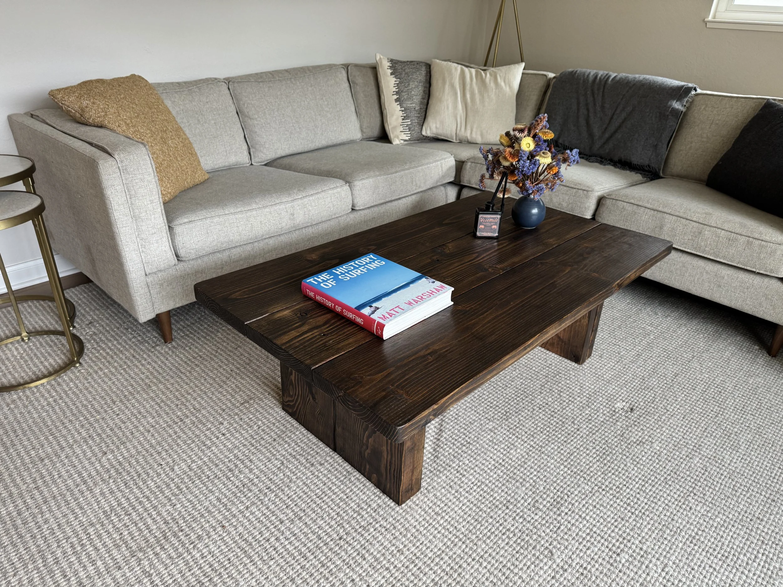 Living room with beige sectional sofa, multiple throw pillows, a dark wooden coffee table with a book and a flower arrangement, a side table, and a floor lamp with a gold stand in the background.