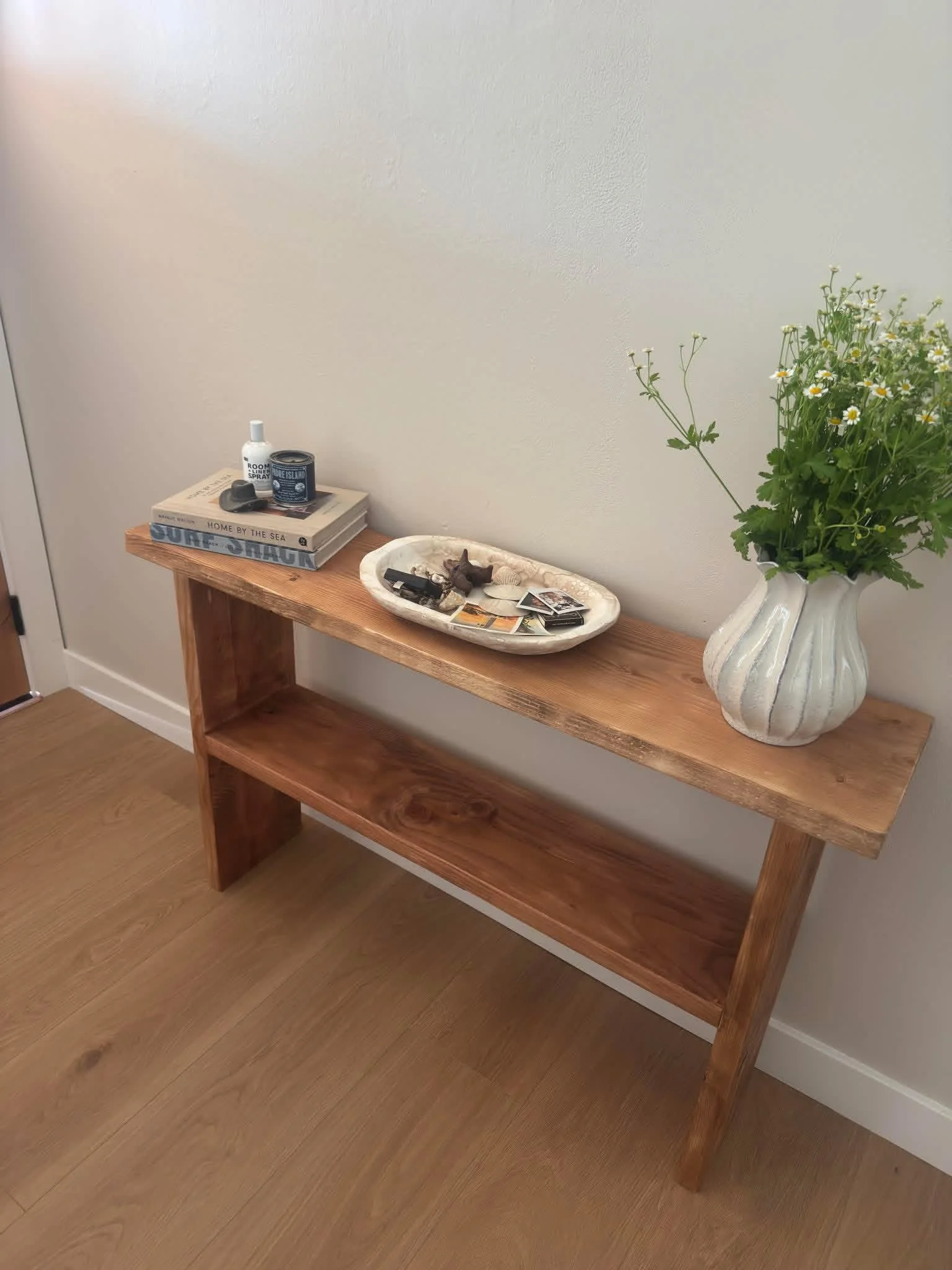 A wooden console table with a flower vase, books, a small tray with decorative items, and spray bottles against a beige wall and wooden floor.