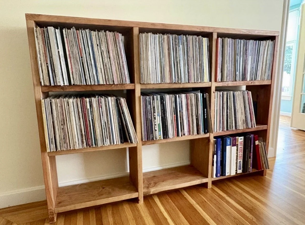 Wooden bookshelf filled with vinyl records and magazines, positioned against a wall in a room with hardwood floors.
