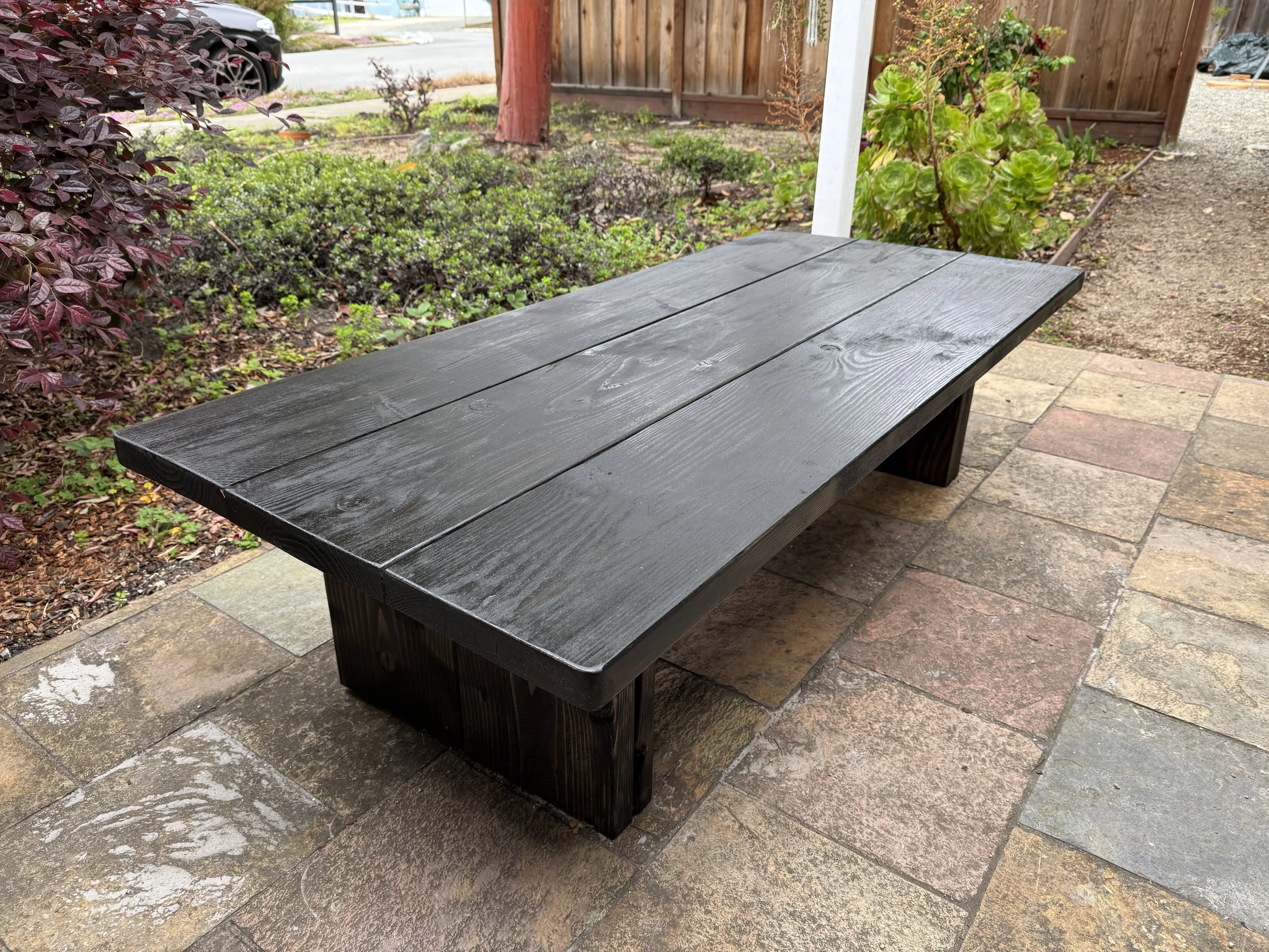 A black wooden outdoor table on a brick patio, surrounded by plants and a wooden fence.