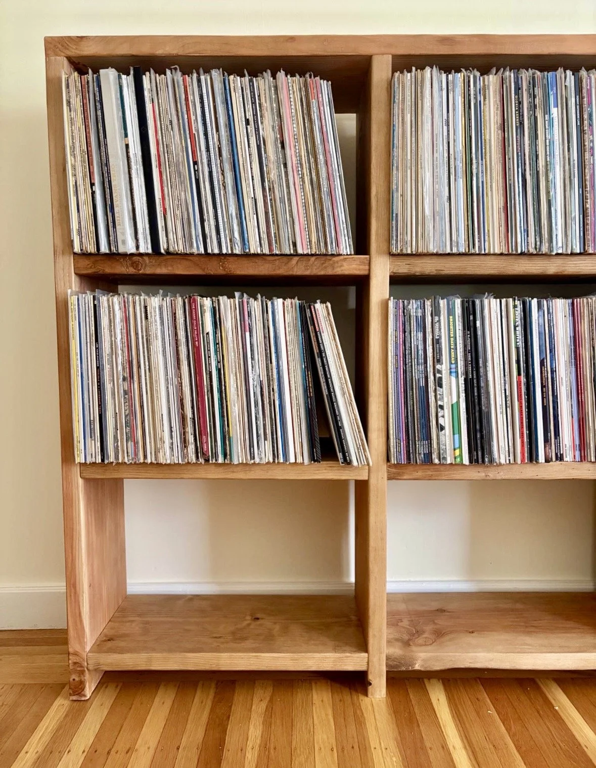 A wooden bookshelf filled with vinyl records, with some records leaning and others neatly arranged, standing on a wooden floor against a light-colored wall.