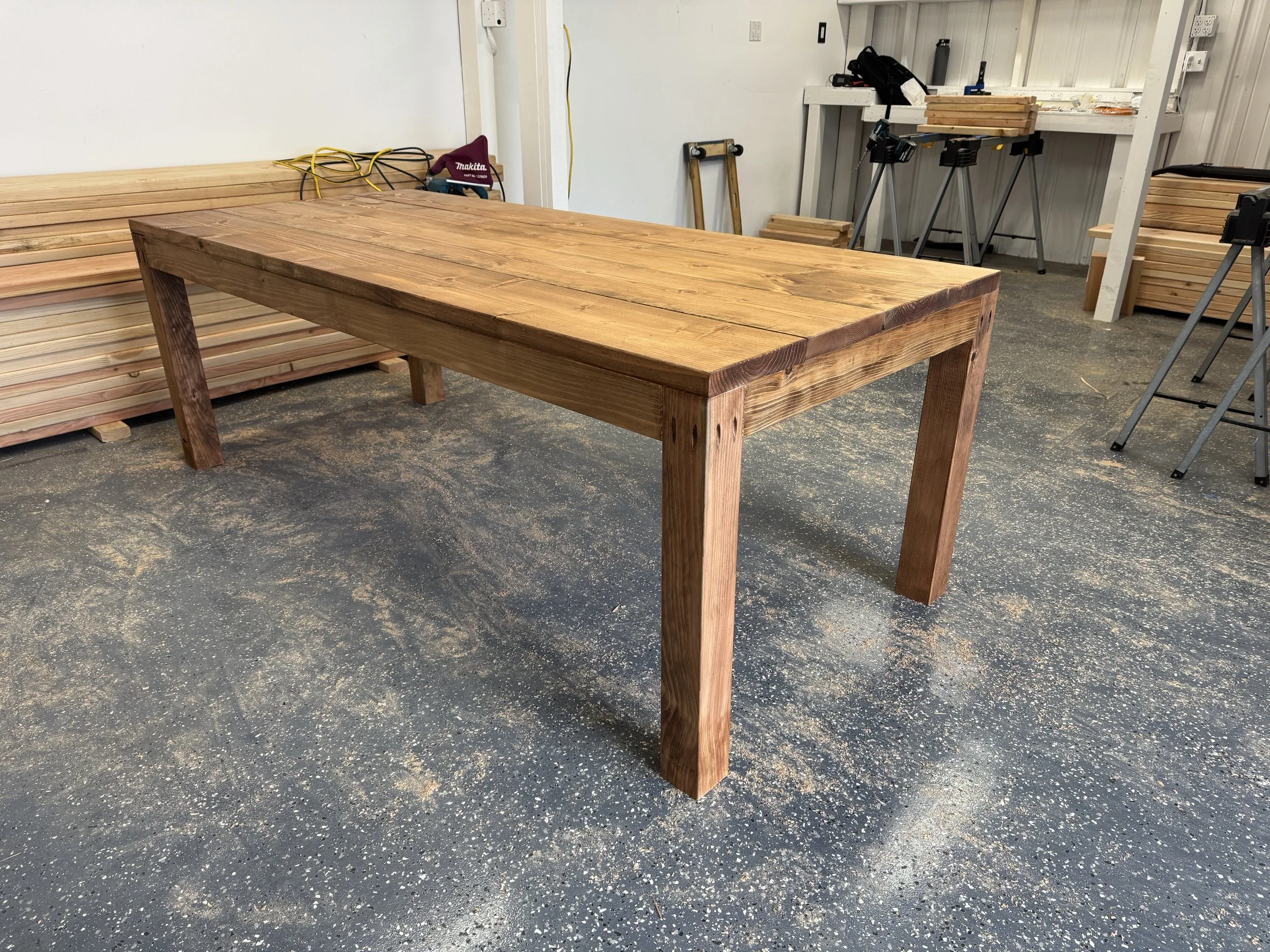 A woodworking workshop with a large wooden table, stacks of cut lumber, and tools such as saws and power drills, with a concrete floor and white walls.