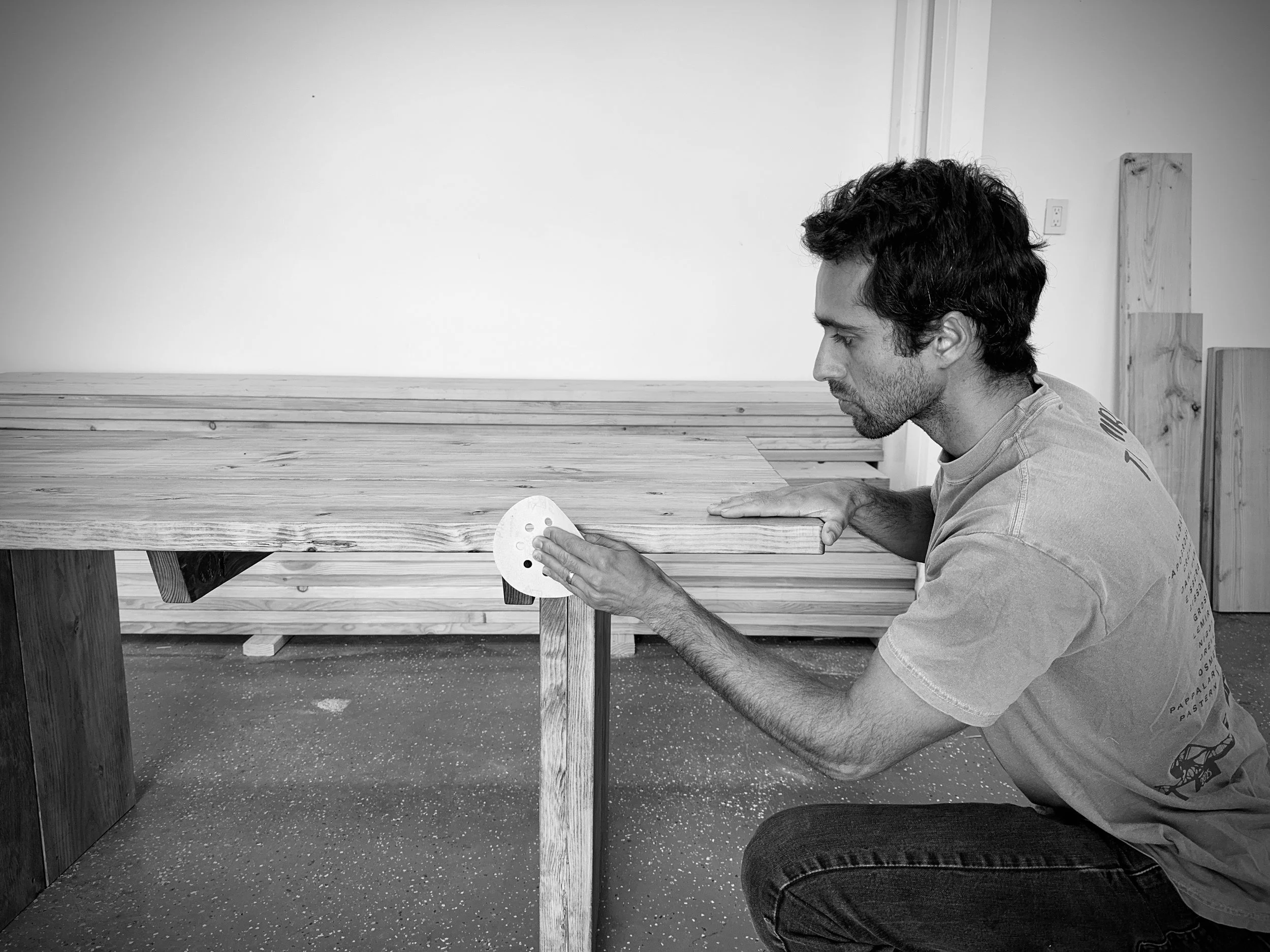 A man is kneeling and sanding the edge of a wooden table in a room with white walls and a dark floor.