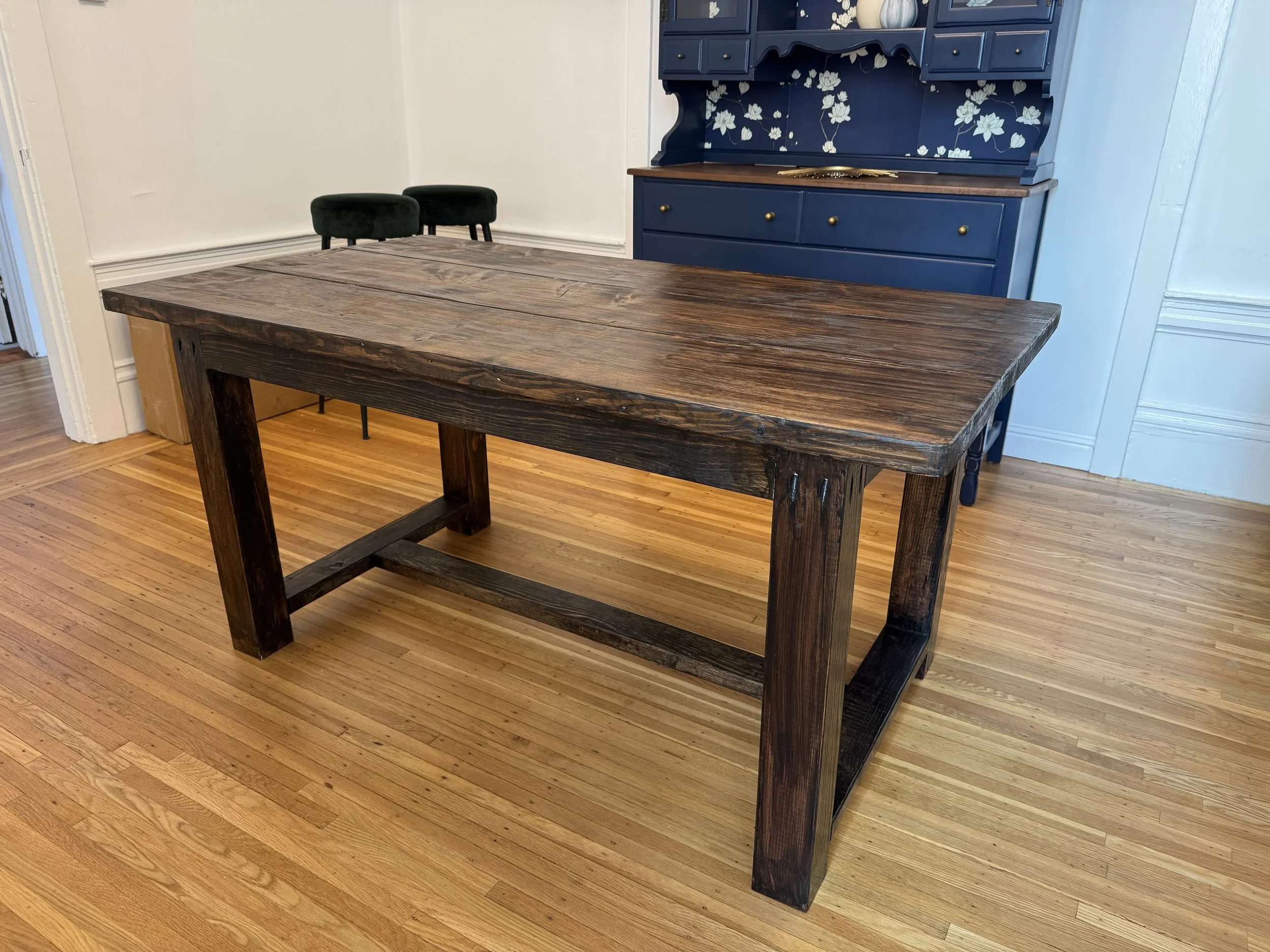 A wooden dining table with a dark, rustic finish in a room with light-colored walls and hardwood flooring. In the background, there is a blue hutch with floral patterns and brass knobs, and two dark green upholstered chairs tucked into the corner.