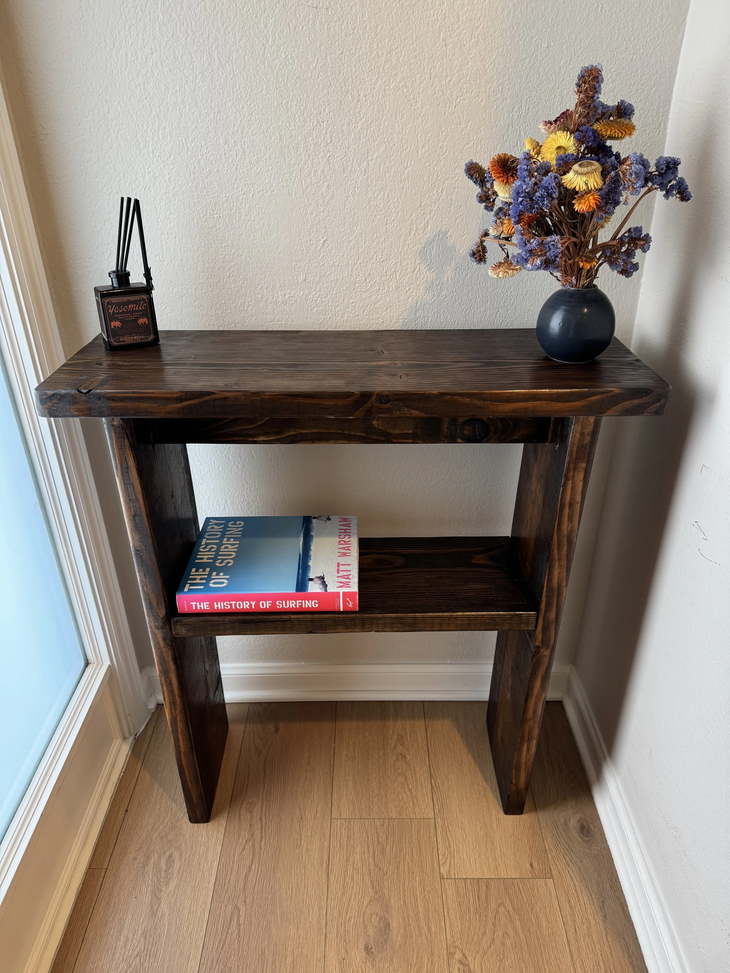 A dark wooden console table against a beige wall, with a black vase containing dried blue and yellow flowers on the right and a book titled 'The History of Surfing' on the bottom shelf.