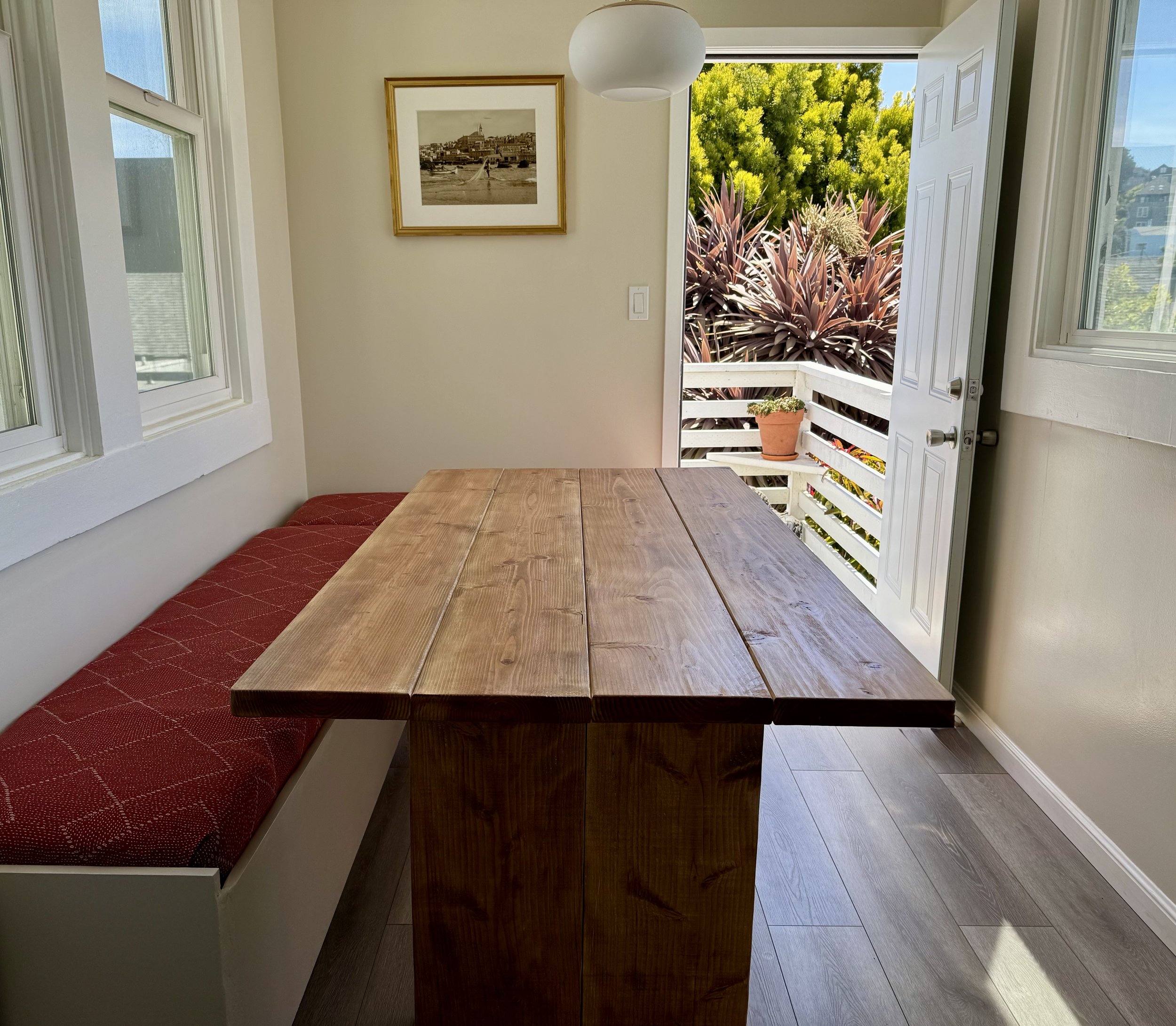 Indoor dining area featuring a wooden table, a red cushioned bench, and windows with a door open to a balcony with potted plants. There is a framed picture on the wall and a round light fixture hanging from the ceiling.