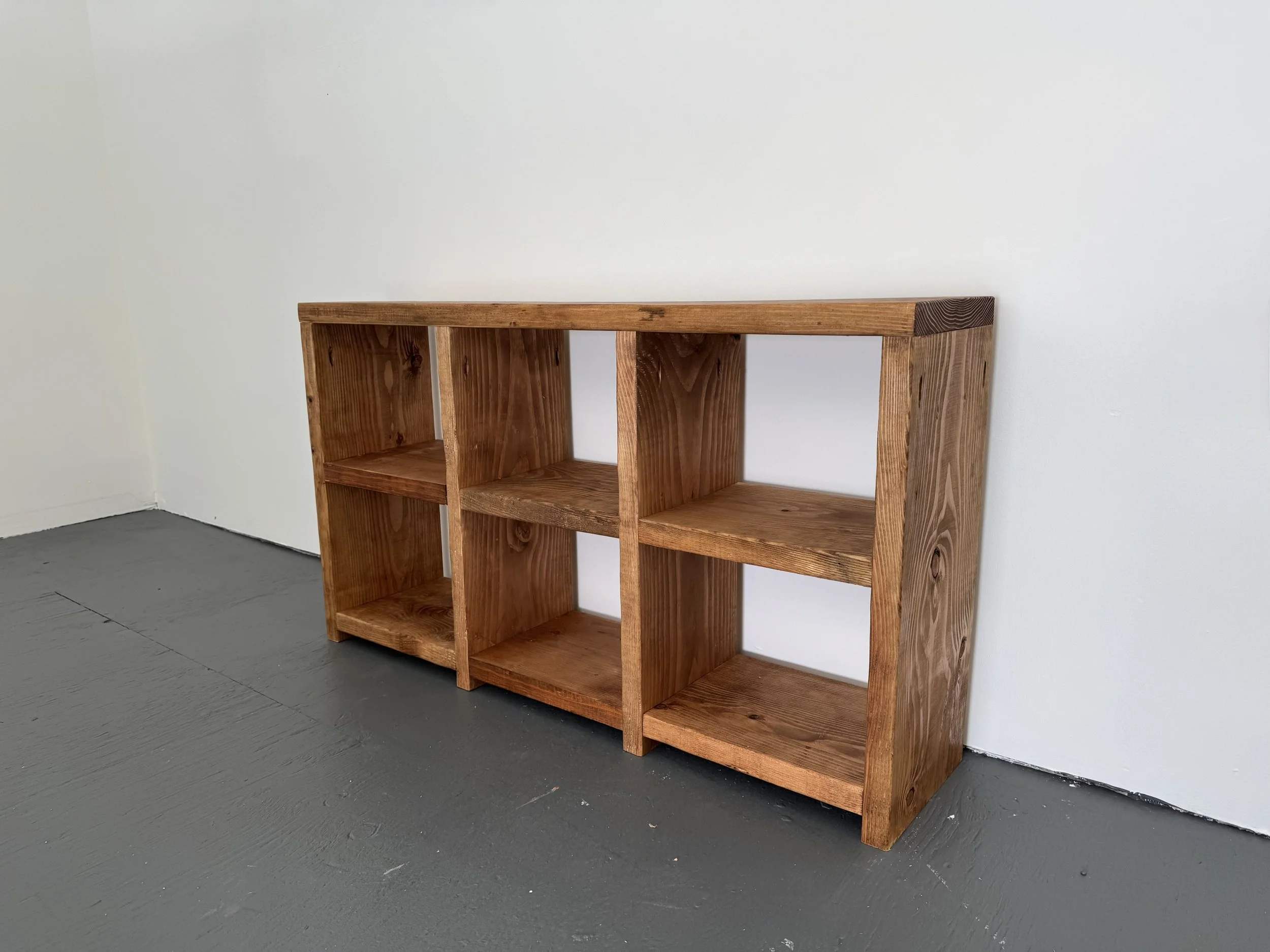 A wooden bookshelf with multiple compartments against a plain white wall, situated on a dark gray floor.