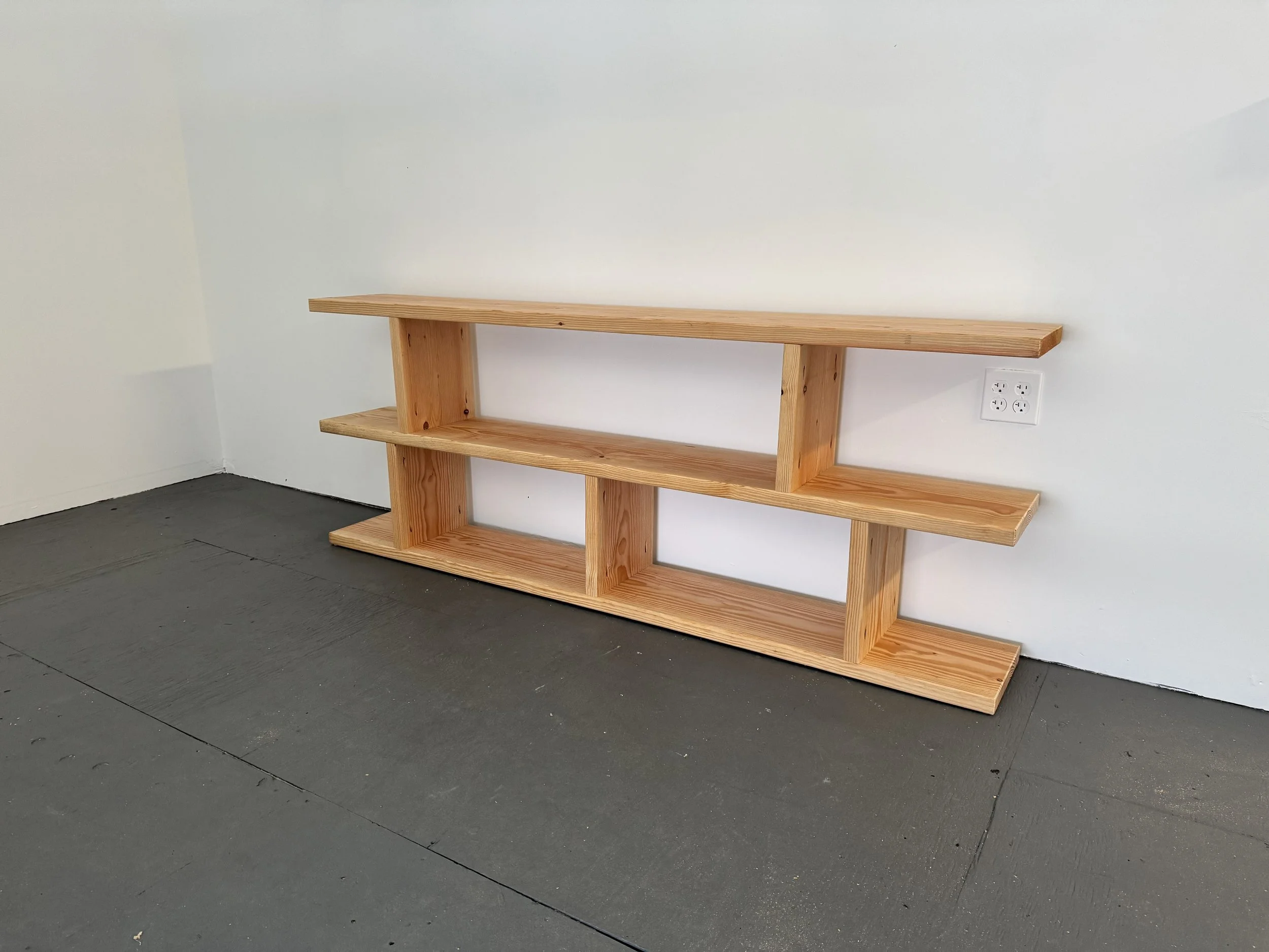 A wooden shelf with two levels secured to a white wall with an electric outlet visible below it, on a dark flooring.