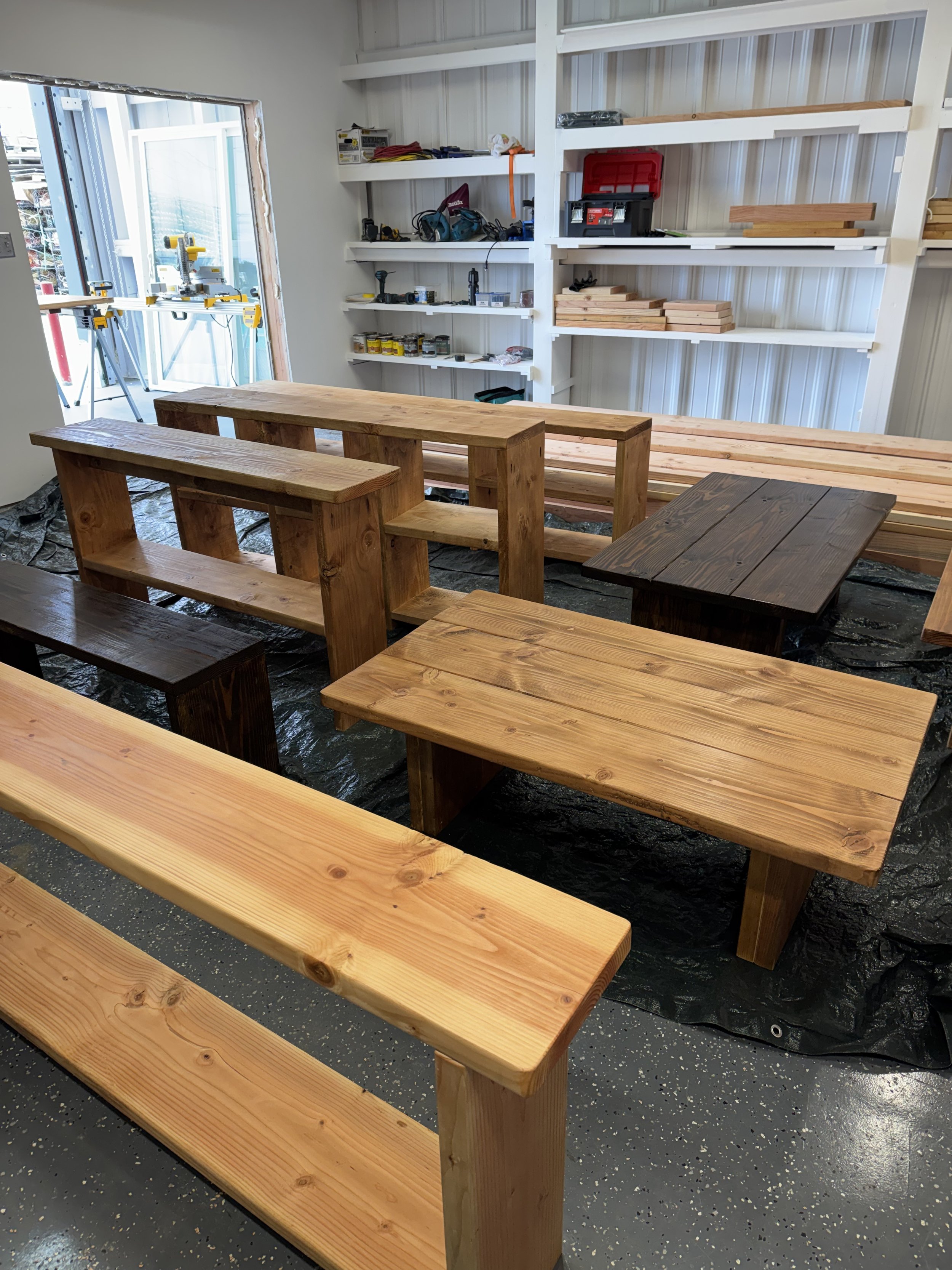 Wooden tables and benches in a workshop or garage, with shelving and tools visible in the background.