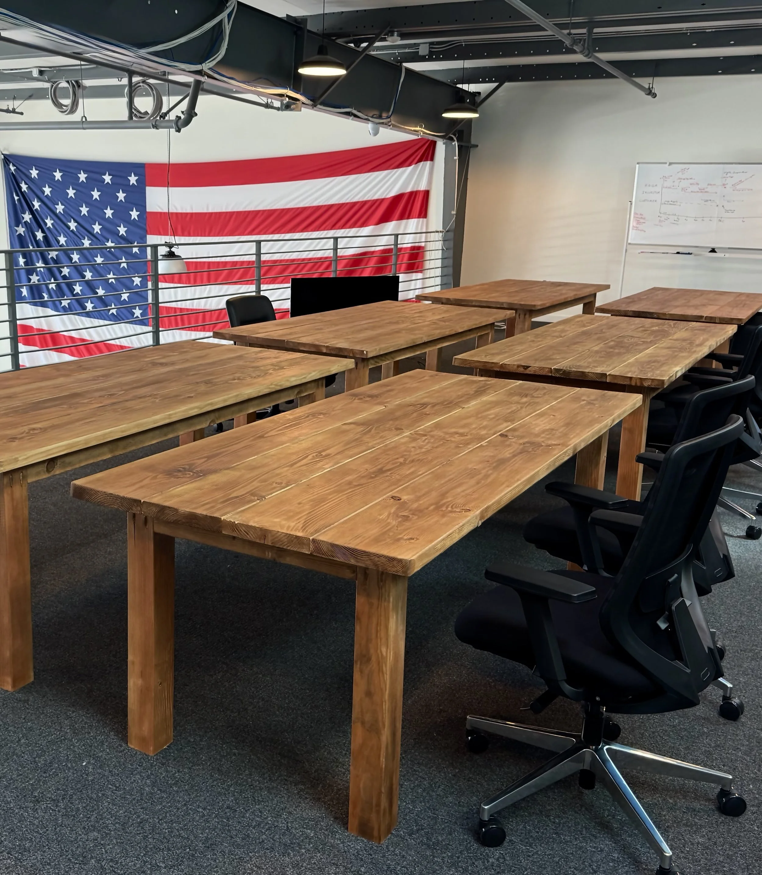Office conference room with a large American flag wall hanging, wooden tables, black ergonomic office chairs, a whiteboard with writing, and exposed industrial ceiling with lighting and pipes.
