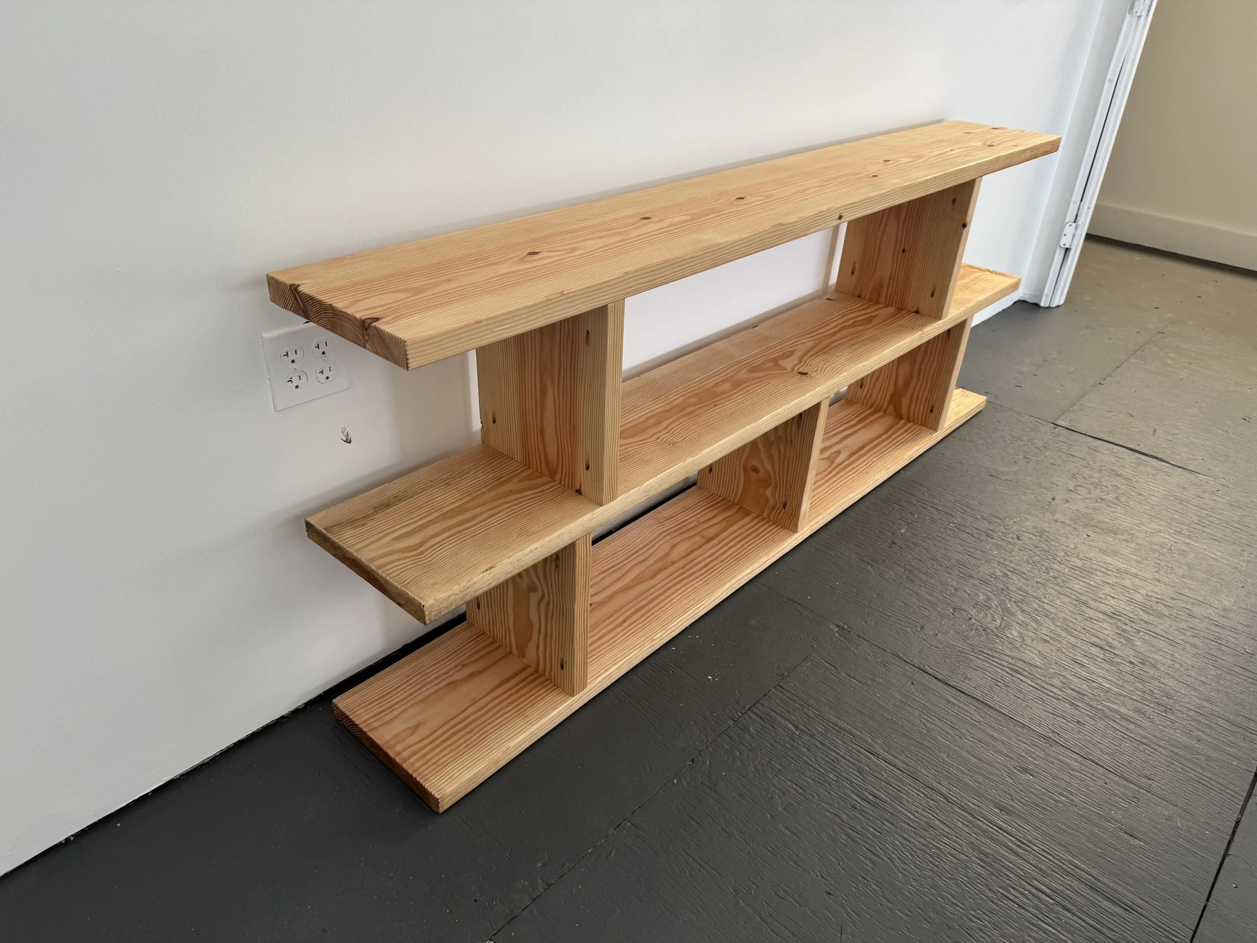 A multi-tier wooden bookshelf made of natural, unfinished wood, positioned against a white wall and placed on a dark floor.