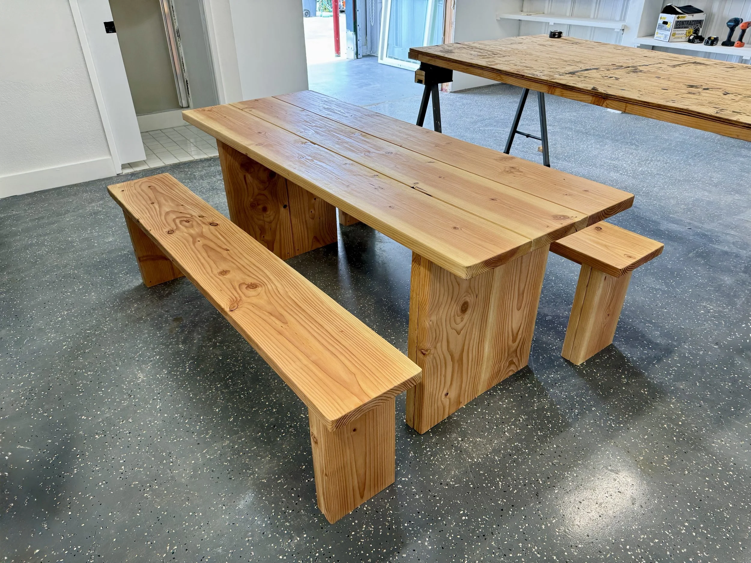 Interior of a workshop with a large wooden picnic table and a bench made of wood. There is a table saw in the background with various power tools on a shelf.
