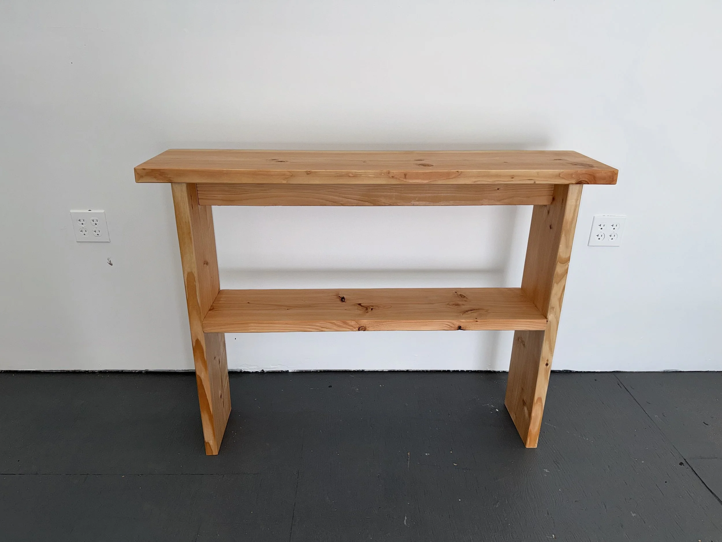 A wooden table with a rectangular top and four angled legs against a plain white wall with two electrical outlets, on a dark floor.