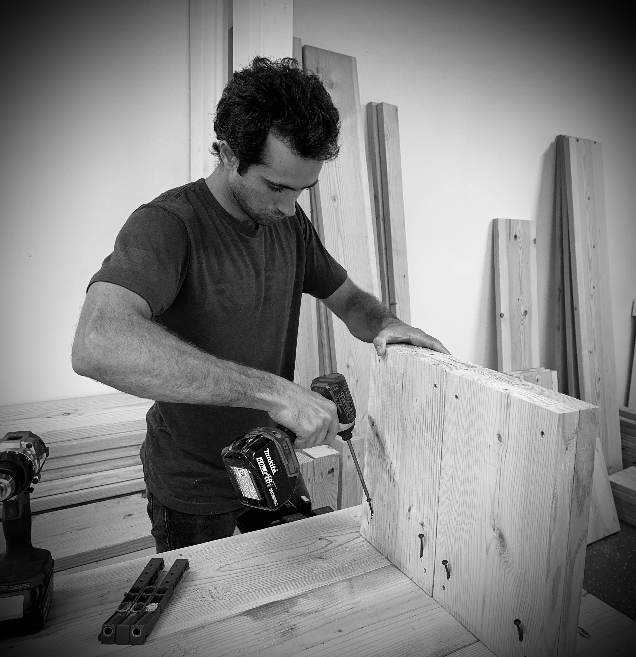 A man working with power tools and wooden planks in a woodworking workshop.