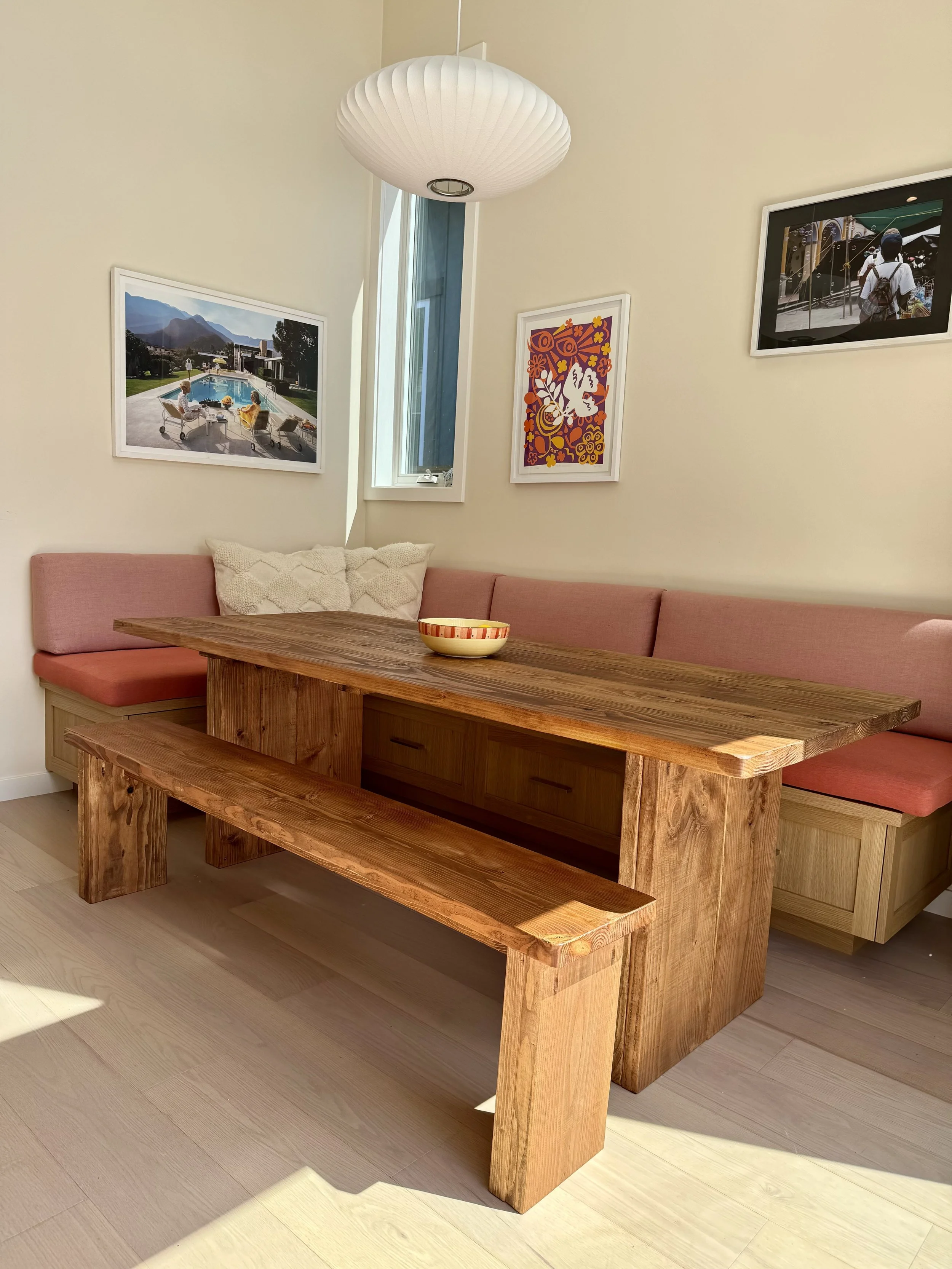 A wooden dining table with a matching wooden bench on one side and peach-colored upholstered seating along the wall. A bowl is placed on the table. The background features framed photos on the wall, a window, and a white paper lantern ceiling light.