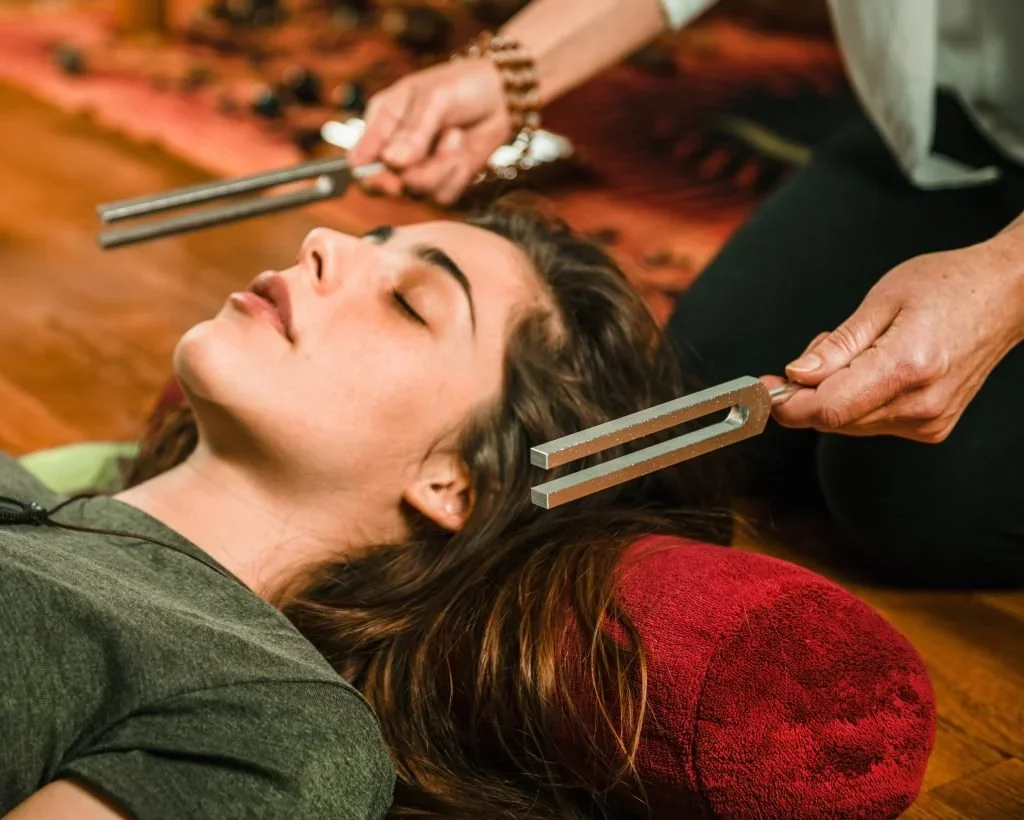 A woman lying on her back with her eyes closed, receiving an sound healing or tuning fork therapy with two metal tuning forks held near her head. The practitioner is partially visible, holding the tuning forks in both hands.
