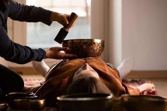 A person receiving a sound healing or therapy session with a singing bowl placed on their chest, while another person holds a hammer or mallet above the bowl.