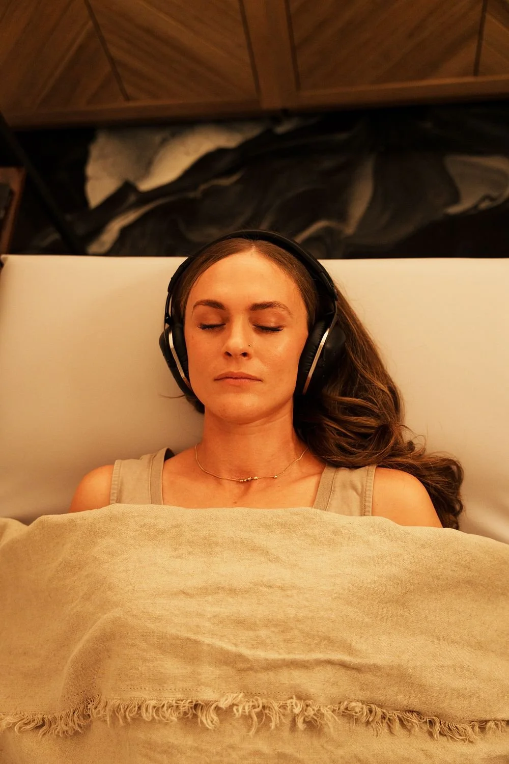 A woman with long brown hair, wearing headphones, lying on a bed with eyes closed, covered with a beige blanket.