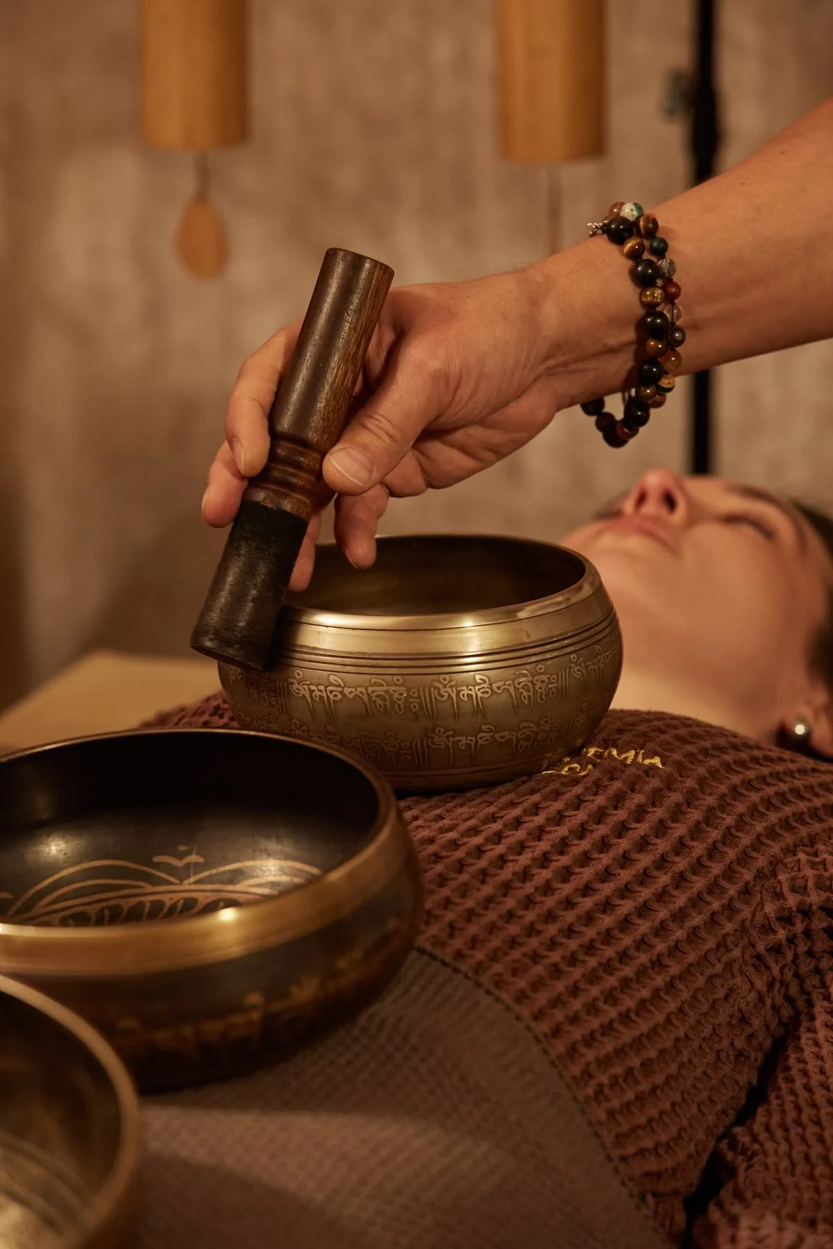 Person laying on a bed with a maroon blanket, receiving a sound therapy treatment with singing bowls, a wooden mallet, and a beaded bracelet on their arm.