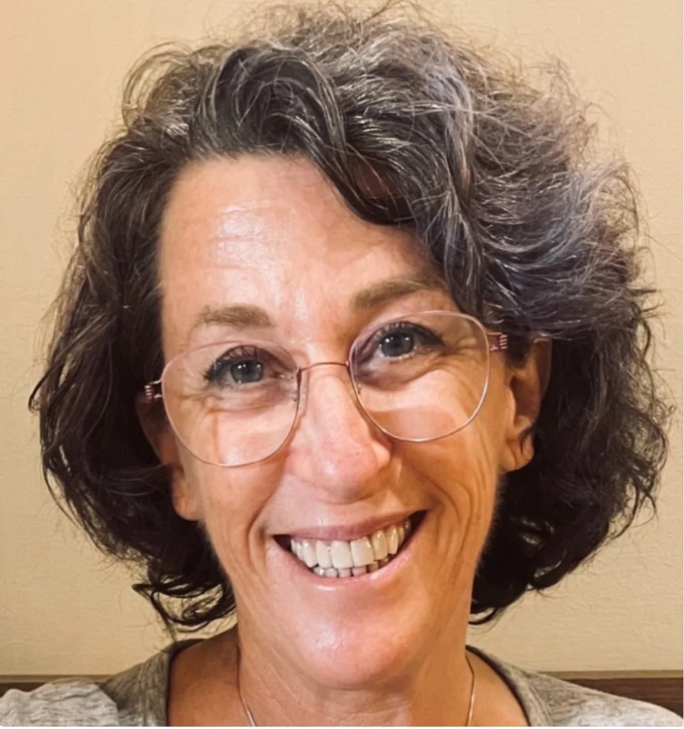 Close-up of a smiling woman with curly brown hair, glasses, and light skin.