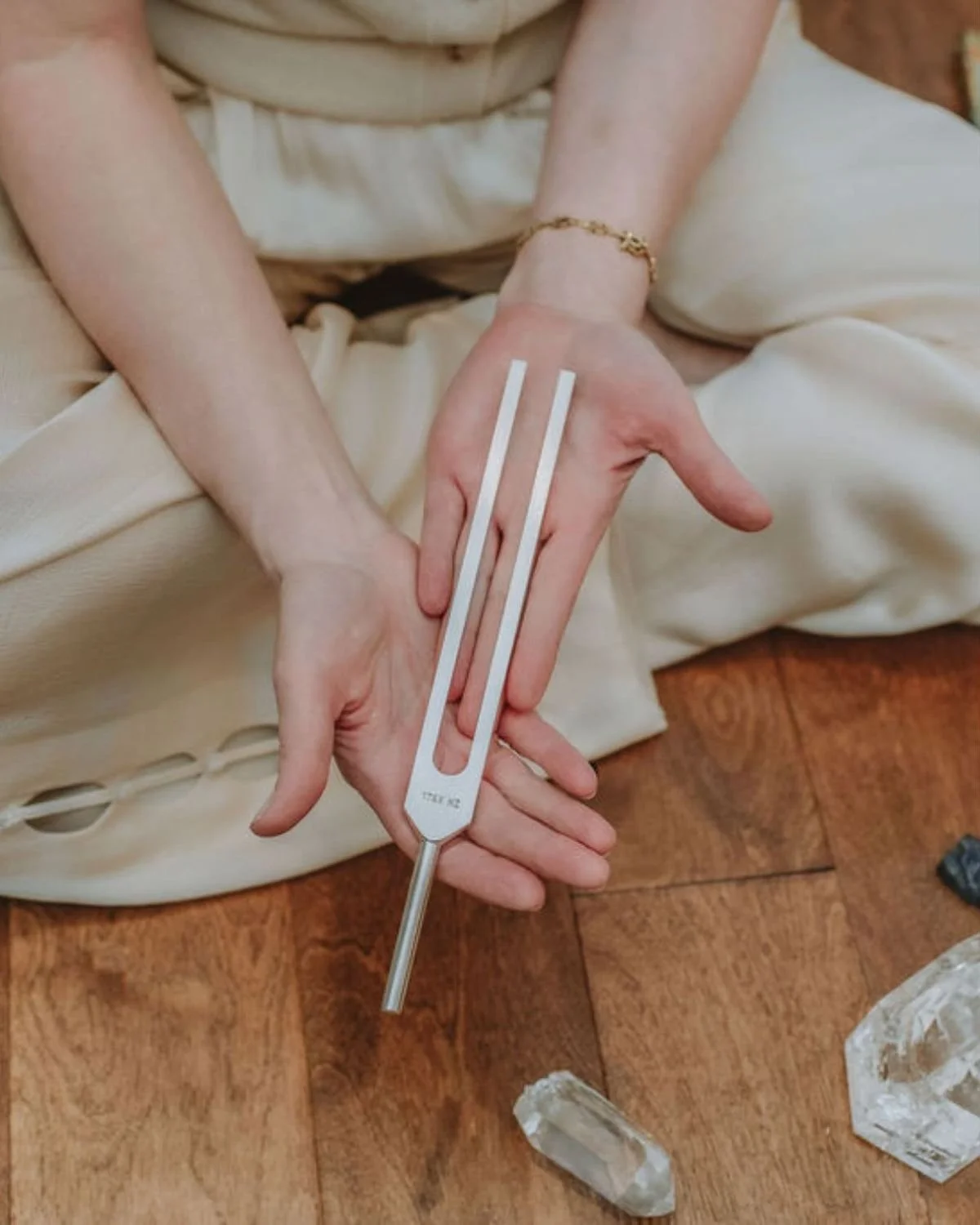 Person holding a tuning fork with two parallel white lines on it, sitting on a wooden floor with large white crystals nearby.