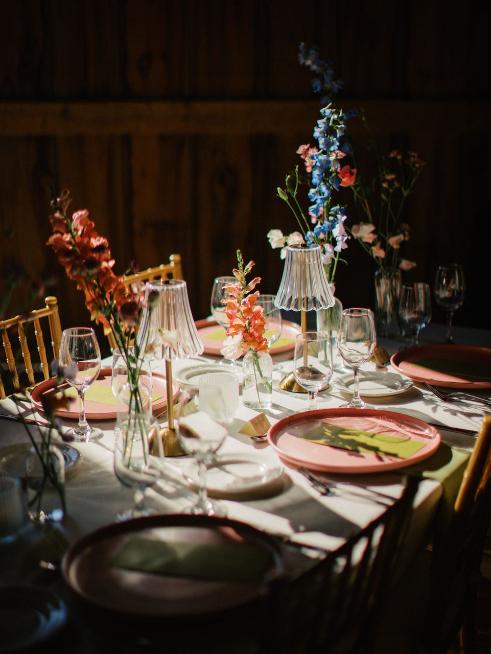 A table set for a meal with pink and white plates, wine glasses, decorative lamps, and vases of pink and blue flowers, in a dimly lit room.
