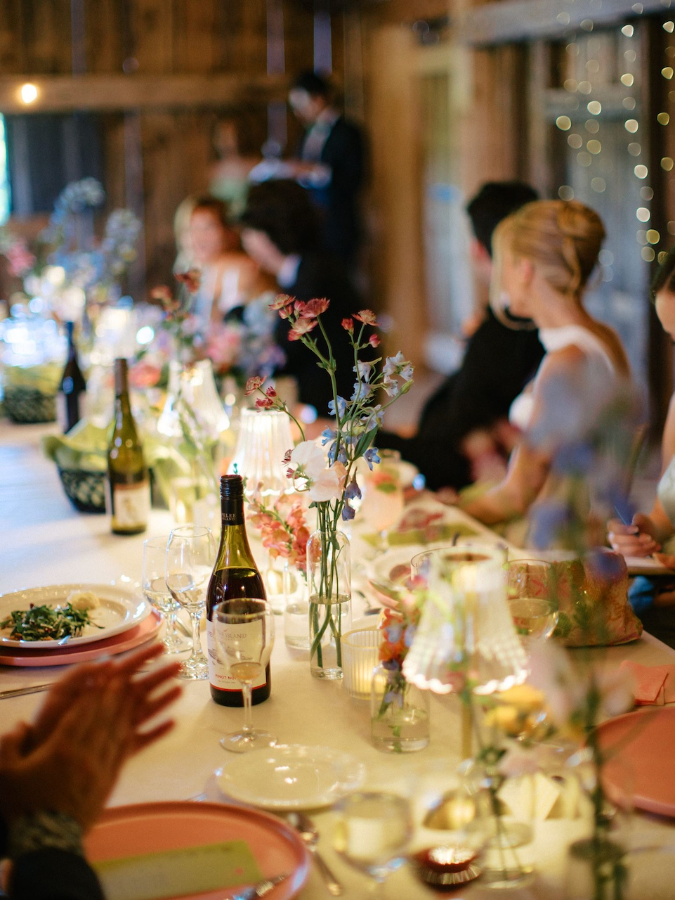 A decorated wedding or celebratory dinner table in a rustic wooden barn with floral centerpieces, glasses, wine bottles, and candles, with people seated and a person standing in the background.