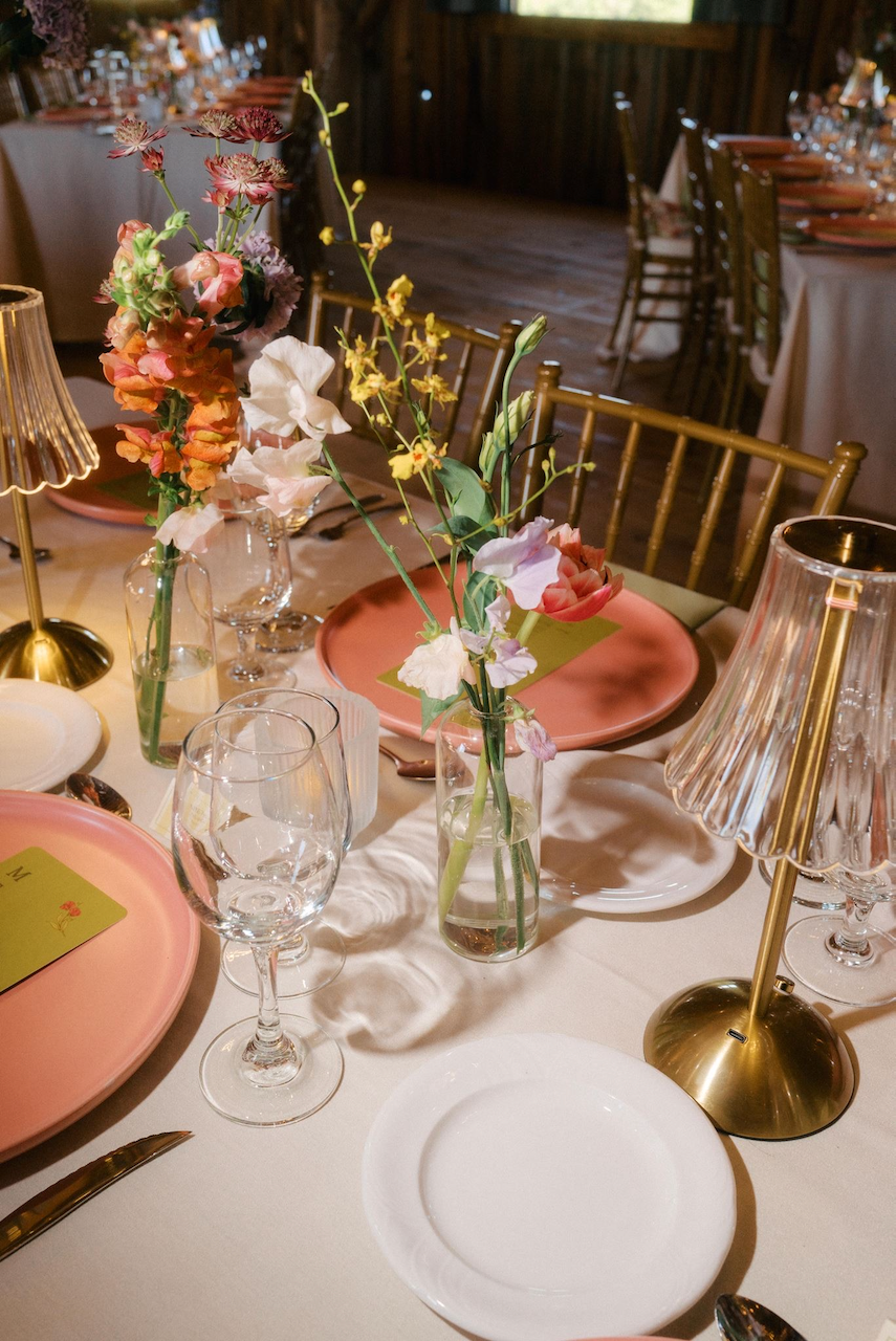 A decorated dining table with pink plates, clear wine glasses, white plates, wrapped utensils, and floral centerpieces in glass vases. Gold lamp bases with pleated shades are also on the table.