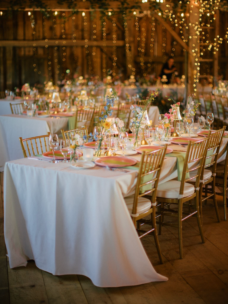 Wedding or event reception setup in a rustic barn with string lights, decorated tables with pink and yellow floral centerpieces, candles, glassware, and gold chairs.