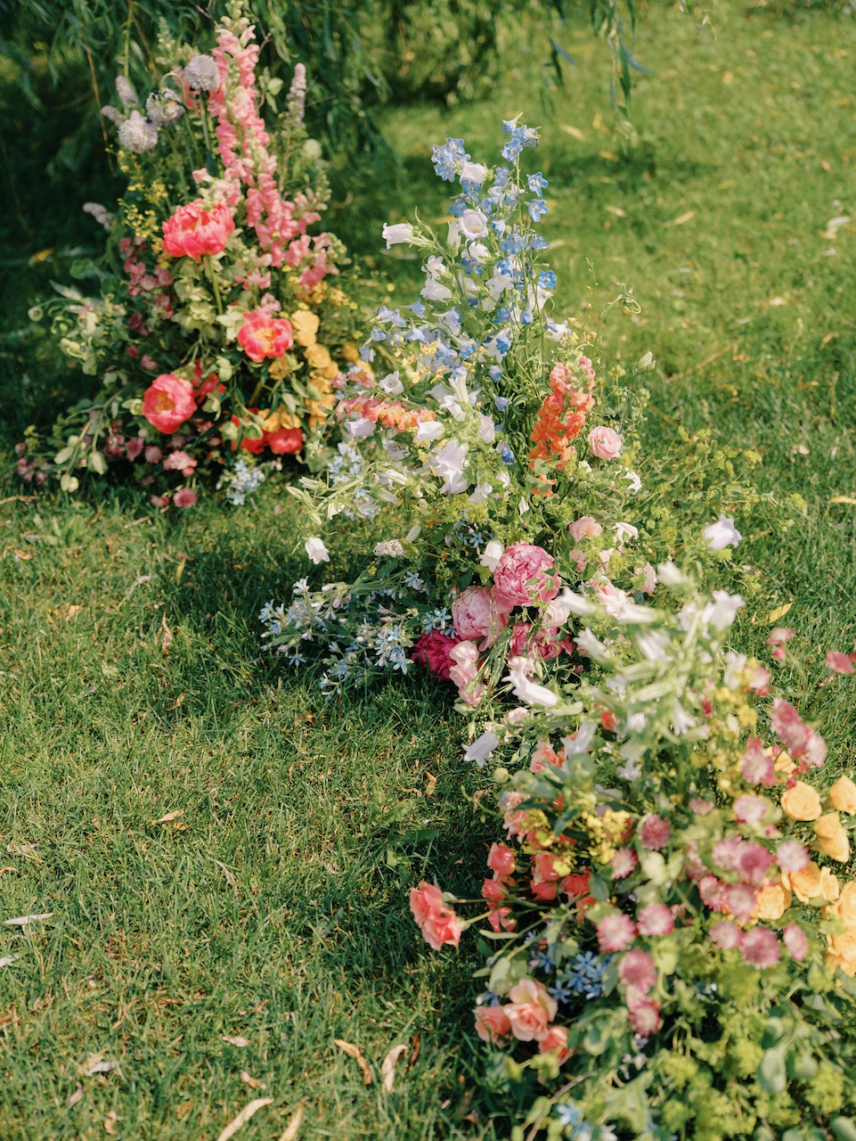 A whimsical pastel floral arrangement on a grassy lawn with various pink, yellow, and blue wildflowers.