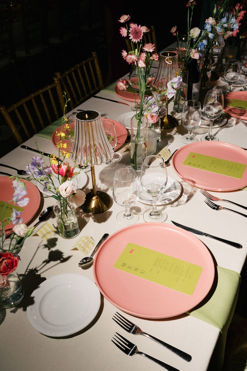 A decorated dining table with pink plates, wine glasses, floral centerpieces, and small lamps.