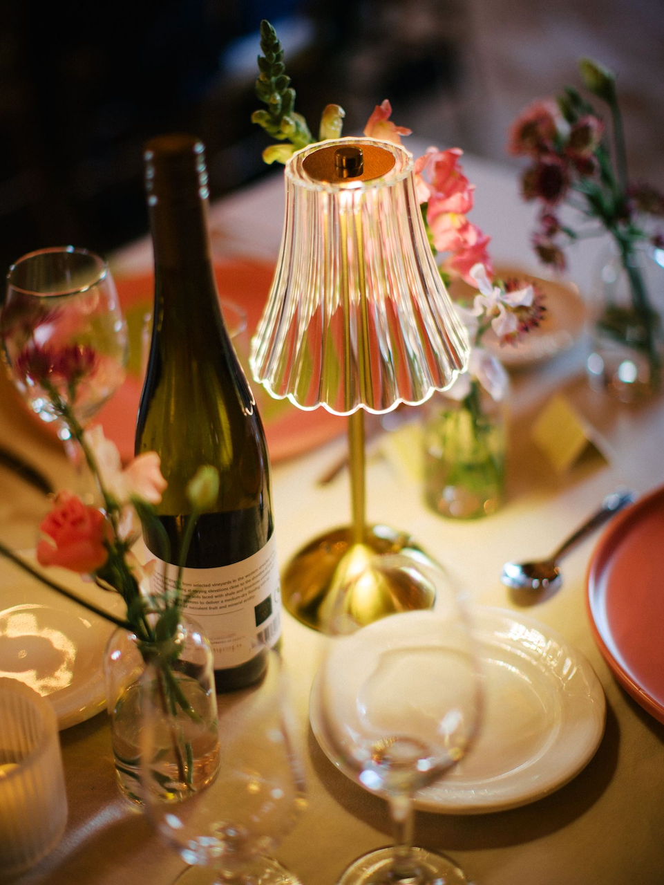 Table setting with a decorative table lamp, wine bottle, wine glass, pink flowers in vases, white plate, pink bowl, and silver spoon.